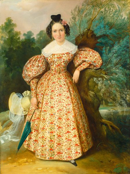This painting shows a woman standing next to a tree, her left arm resting on a low branch. She has fair skin, rosy cheeks, large eyes, and dark curled hair in an updo and adorned with a pink flower. She wears a dress with a red-and-green floral pattern on a pale-yellow background, puffed sleeves, a lacy collar, and a wide belt. She is holding a closed green umbrella in her right hand. A pale-yellow hat with a veil rests on the ground behind her to the left. She stands on a brown patch of dirt, and to the right appears to be a body of water with green reeds. The background shows dense green trees under a sky of pastel pink clouds, a small patch of blue visible near the top left.