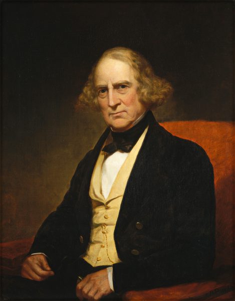 The image shows the upper half of a seated man. He is positioned slightly angled to the right and looking directly at the viewer. The man has a stern expression, characterized by his prominent brow and deep-set eyes. His hair is light brown with some natural curl, extending just below his ears. He is dressed in formal attire, including a dark coat, a cream-colored waistcoat, a white shirt, and a black cravat. His hands rest on his lap, one hand holding a pair of spectacles. A gold chain hangs from a pocket of his waistcoat. The background is a dark, muted shade that contrasts with the warm tones of the chair and his clothing.