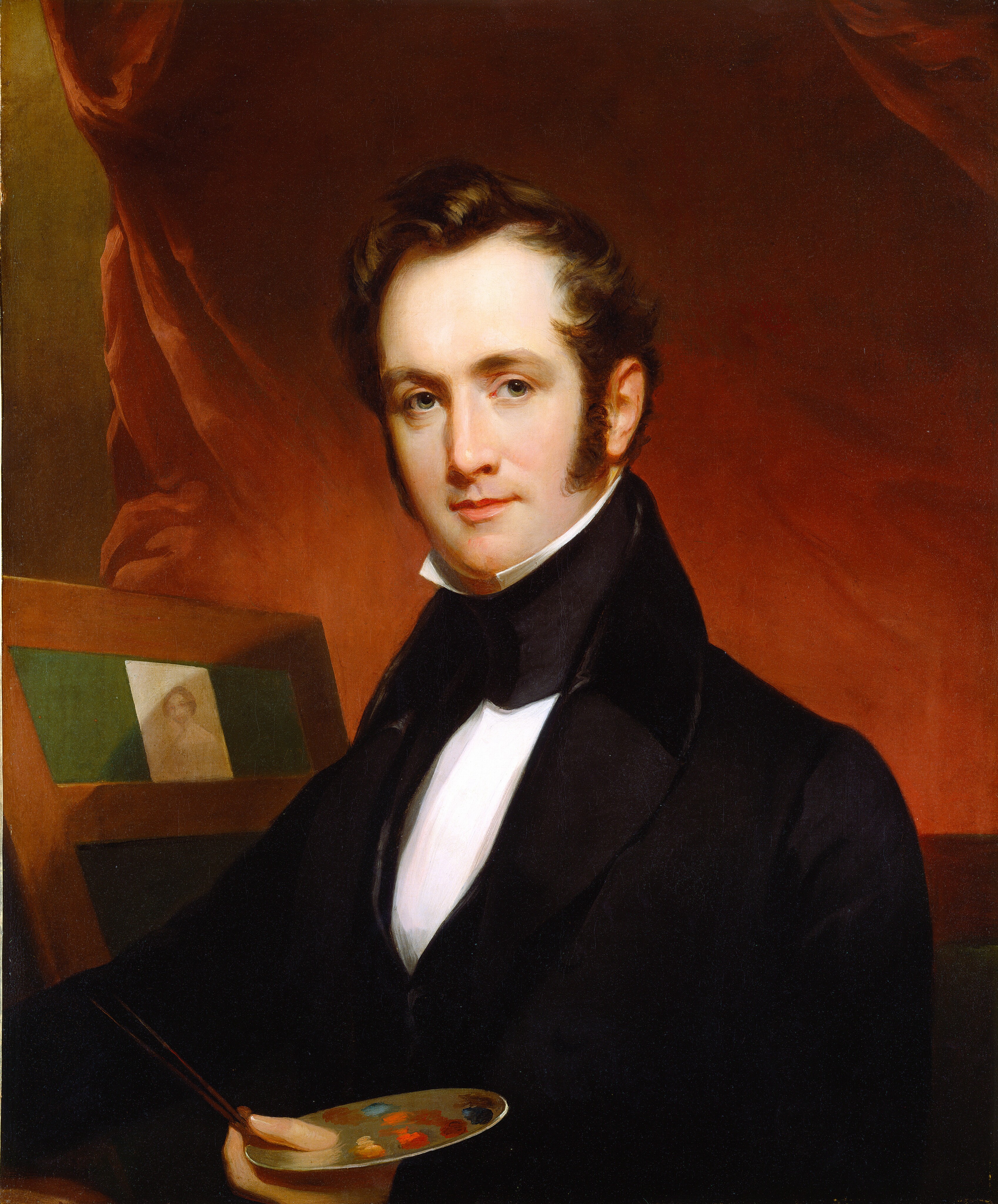 The image shows a man seated, turned slightly to the left, looking directly at the viewer. He has fair skin, light-colored eyes, and dark brown wavy hair with sideburns reaching his jawline. He wears a black coat with a high collar over a white shirt. He sits near an easel with a sketch visible, holding a palette and paintbrushes in his left hand. The background has draped fabric in red or brown shades.