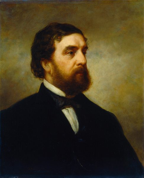 The image is a portrait of a man shown from the shoulders upward. He is in a side profile facing to his right. The man has a full beard and mustache, brown, slightly curly hair, a broad forehead, and deep-set eyes. He wears a dark jacket, white shirt, and a black bow tie. The background is a blend of brown and grey tones.