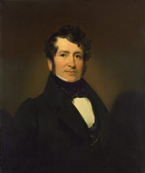 The image shows a man's upper body in a formal portrait setting. He is depicted sitting or standing upright with his head turned slightly to the right, directly facing the viewer. The man has fair skin, distinct facial features including a full face with a slight smile and prominent cheeks. His hair is dark, thick, and curly, framing his face neatly. He is dressed in formal attire, wearing a dark coat and a white shirt with a high collar, complemented by a black bow tie. There is a hint of a gold chain peeking from beneath his coat. The background is a plain, dark brown.