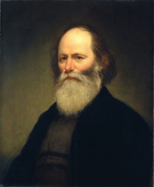 The image is a portrait of a man shown from the torso up. He is positioned slightly turned to the right, facing the viewer with a direct gaze. He has a balding forehead with thin hair at the top, and the remaining hair is long and curly at the sides and back. His facial features include a prominent nose and a full, white beard and mustache that cover most of his lower face. The man is dressed in a dark, subdued garment with a high collar visible around his neck. The background is plain and dark, with subtle variations in color, keeping the focus on the subject. There are no visible jewelry or objects being held.
