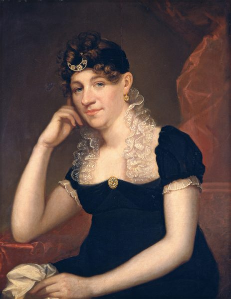 This painting depicts a seated woman from the waist up. She has her right elbow resting on one arm of the red chair that she sits in, with her head leaning on her hand. Her left hand is holding white fabric in her lap. She has light skin, blue eyes, a large nose, and pink lips set in a slight smile. Her curly brown hair is styled up with a decorative headband featuring a crescent-shaped ornament. She wears a dark dress with a lace-trimmed neckline, short, puffed sleeves, a gold brooch, and gold earrings. The background is warm brown with a reddish fabric draped on the right side.