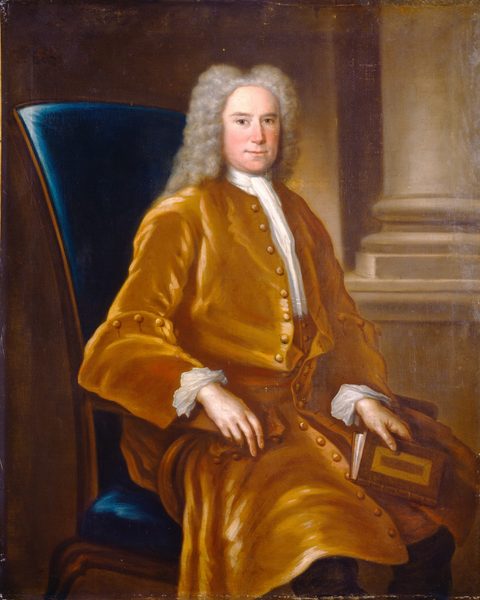 The image shows a man seated in a large blue chair. He is dressed in a golden-brown silk robe, holding a closed book in his lap. The man has a neutral expression and his gaze is directed towards the viewer. He has a broad forehead, light eyebrows, and gray curly hair that falls to his shoulders. The background includes architectural elements like a column on the right side.