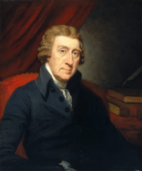 The image shows a man in a dark suit with a white cravat, sitting and facing slightly to his right. He has light brown curly hair, fair skin, light blue eyes, a straight nose, and a subtle smile. The background is a red drapery with books stacked horizontally partially hidden behind it, and the man is illuminated against this backdrop.