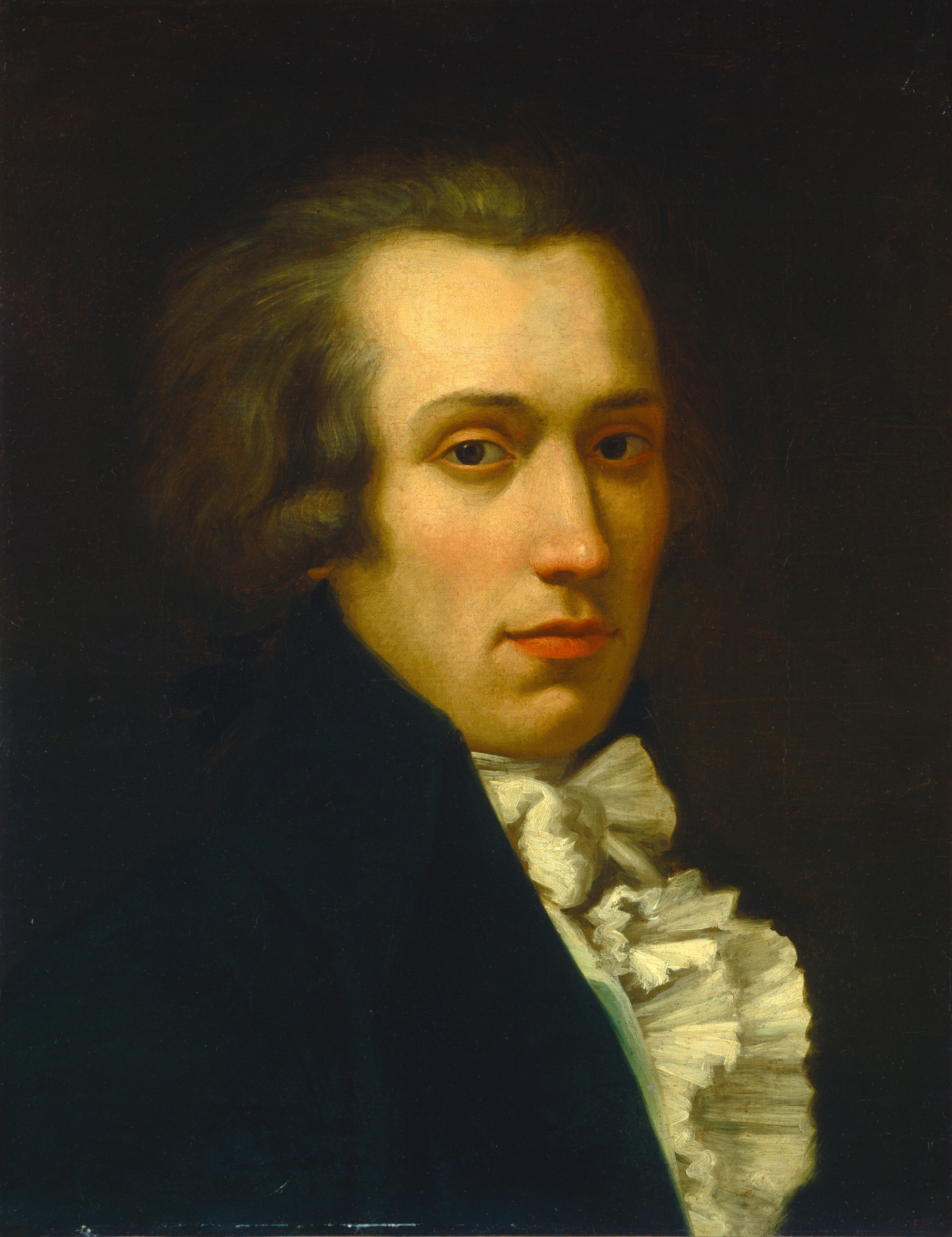 The image displays a chest-up portrait of a person gazing directly at the viewer. The individual has a defined jawline, high cheekbones, and dark, slightly arched eyebrows. Their light brown hair is moderately long and voluminous. They are wearing a dark coat with a high collar and a white ruffled cravat around the neck and upper chest. The background is dark and plain, emphasizing the subject's appearance and clothing.