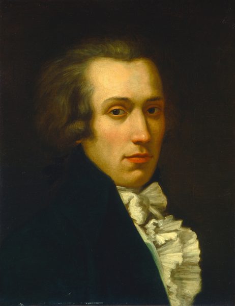 The image displays a chest-up portrait of a person gazing directly at the viewer. The individual has a defined jawline, high cheekbones, and dark, slightly arched eyebrows. Their light brown hair is moderately long and voluminous. They are wearing a dark coat with a high collar and a white ruffled cravat around the neck and upper chest. The background is dark and plain, emphasizing the subject's appearance and clothing.