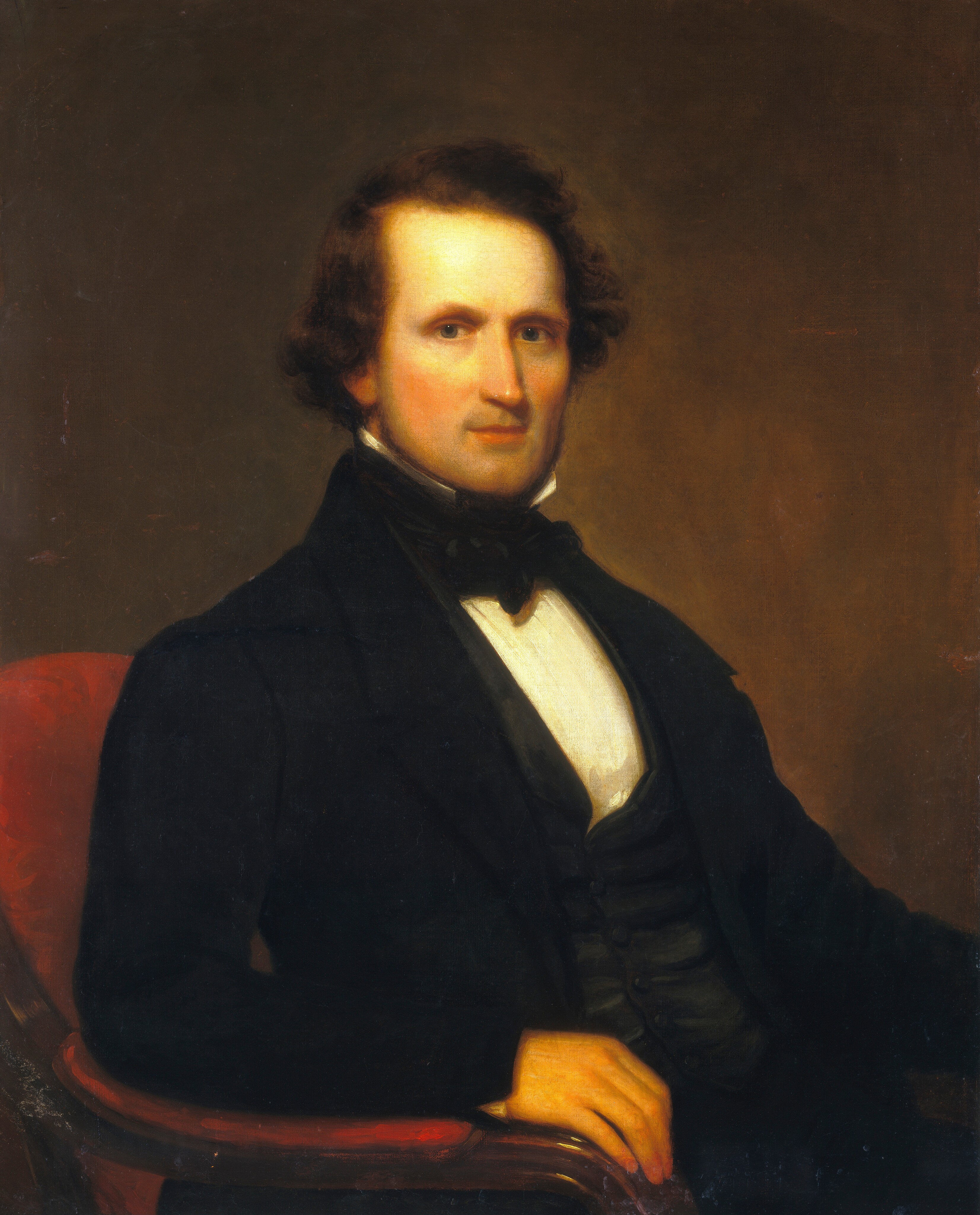 The image is a portrait of a man seated, showing the upper half of his body. He has dark brown hair, fair skin with a warm undertone, a straight nose, thin lips, and pronounced cheekbones. He wears formal attire including a dark suit with a double-breasted waistcoat, a white high-collared shirt, and a black cravat tied in a bow. His right hand rests on the arm of a red upholstered chair. The background is a simple soft brown gradient.