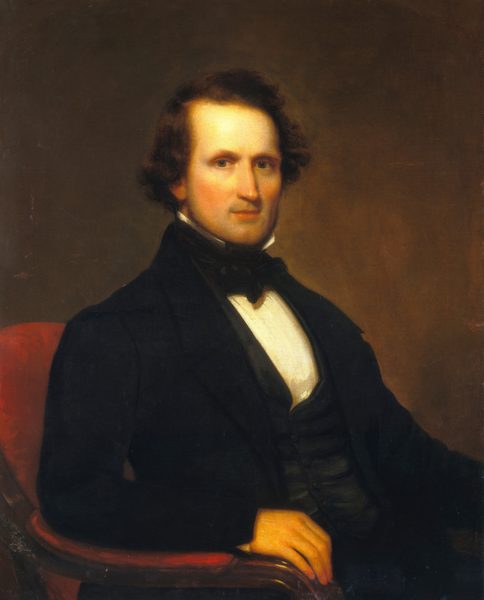 The image is a portrait of a man seated, showing the upper half of his body. He has dark brown hair, fair skin with a warm undertone, a straight nose, thin lips, and pronounced cheekbones. He wears formal attire including a dark suit with a double-breasted waistcoat, a white high-collared shirt, and a black cravat tied in a bow. His right hand rests on the arm of a red upholstered chair. The background is a simple soft brown gradient.