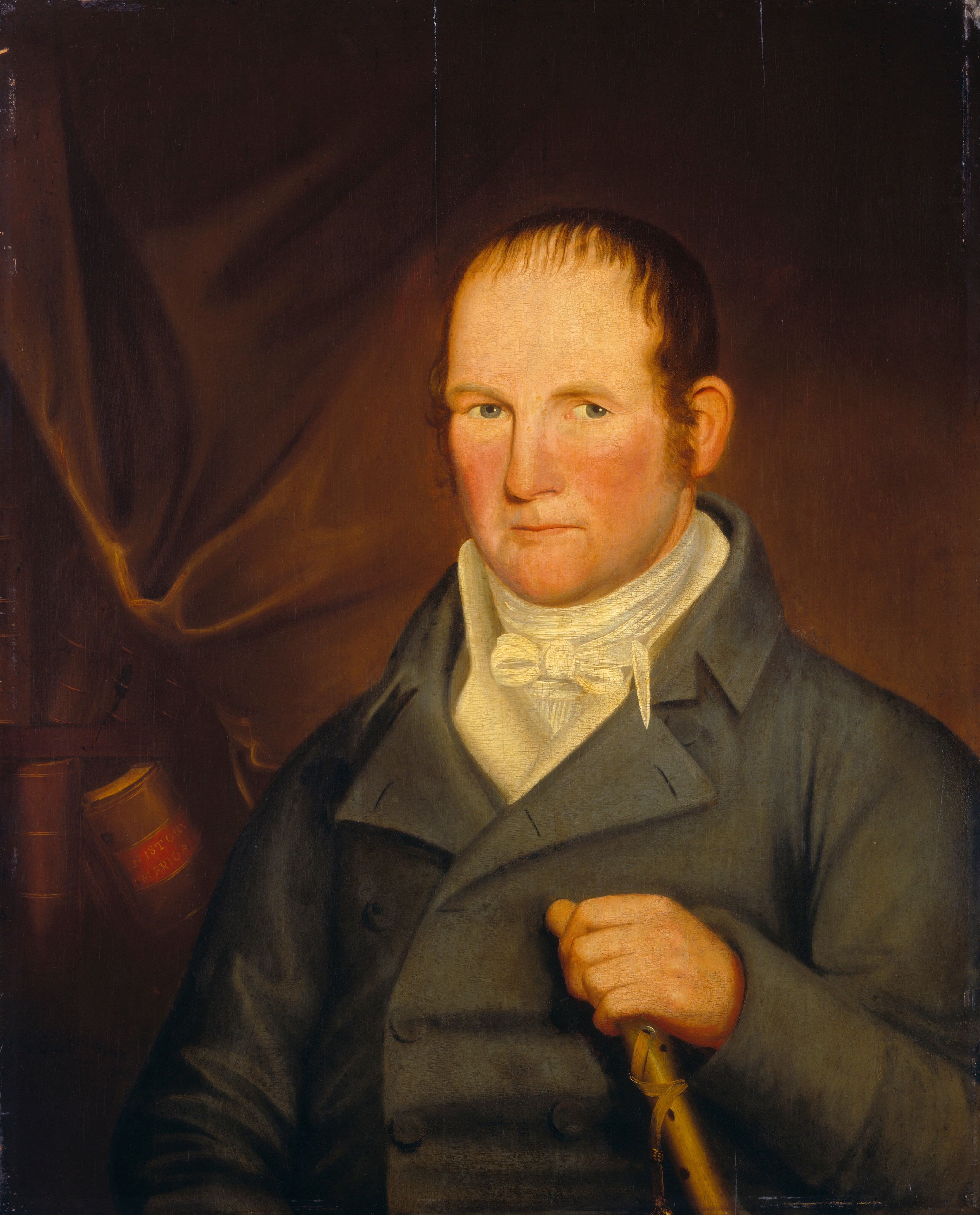 The image depicts a man from the waist up. He stands slightly to the right but faces forward, making direct eye contact. The man has a fair complexion with a reddish hue on his cheeks and a broad face. His hair is short, dark brown, and receding from the forehead. He wears a dark double-breasted coat with large buttons and a white cravat around his neck. His right hand is visible holding onto what appears to be a walking stick or cane. In the background, there is a dark draped cloth on the left side, and beside this, the spine of a book, part of a small bookshelf, can be seen. The overall background is dark, emphasizing the figure in the portrait.