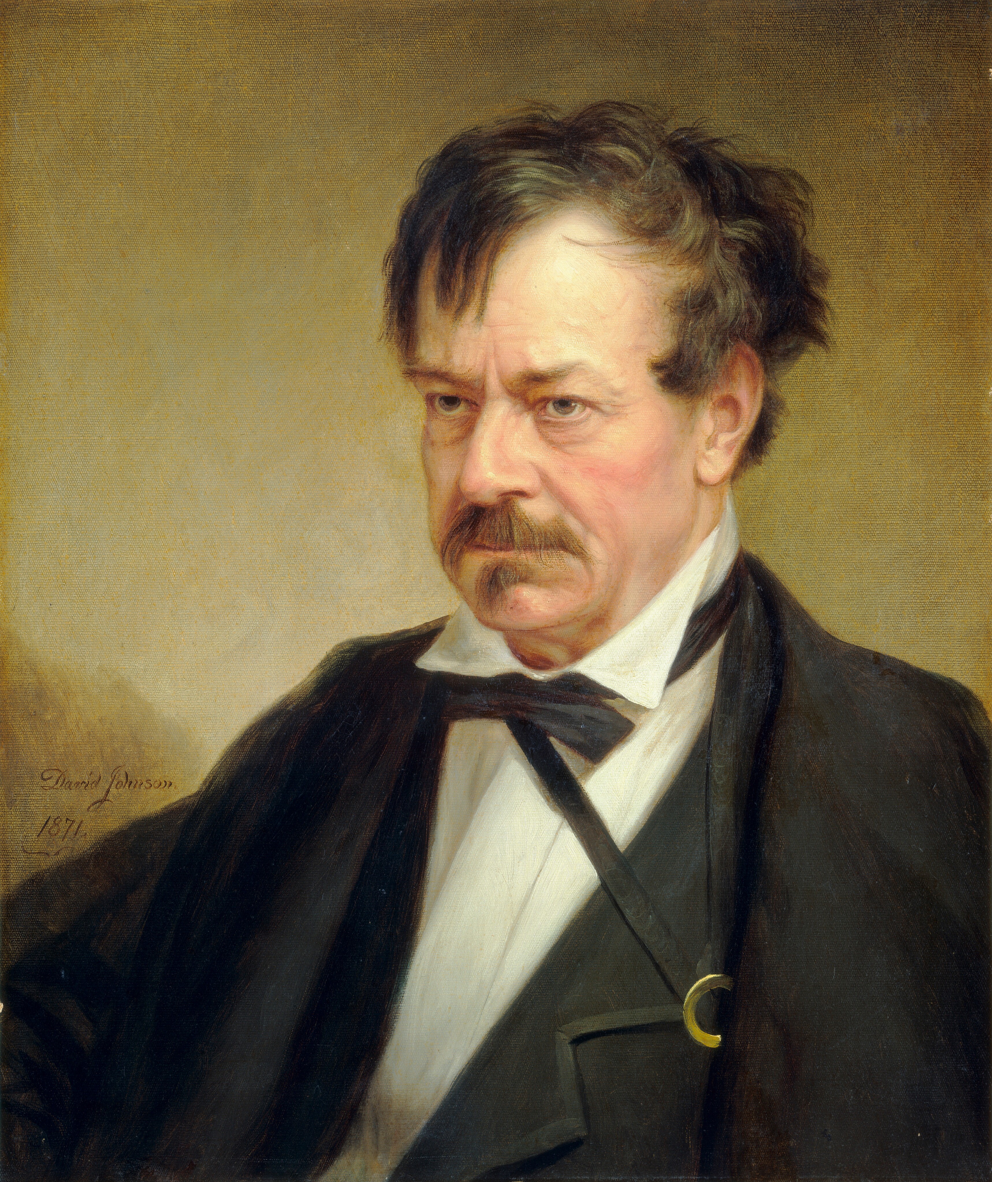 The image is a portrait of a man showing the upper half of his body, positioned slightly turned to one side. He has a serious expression with a prominent mustache and slightly disheveled dark hair that appears to be thinning at the crown. He is dressed in formal attire, wearing a black coat over a white shirt with a high collar and a black bow tie. A gold hoop in the buttonhole of his coat adds a small note of detail. The background is a simple, softly shaded neutral color.