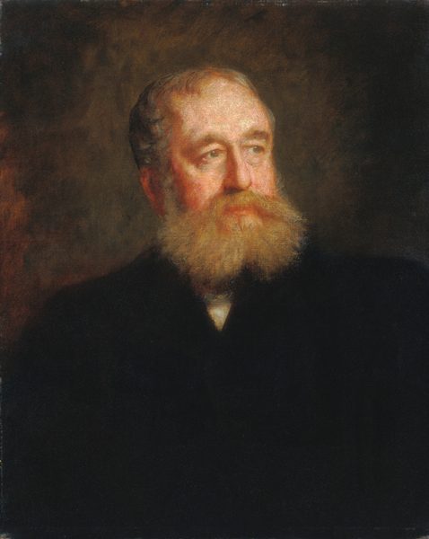 The image shows a portrait of an older man from the shoulders up. He is facing slightly to the right, with his gaze directed off-camera. The man has a large, bushy beard that is a mix of white and reddish tones. He has a receding hairline with short, thinning hair on top. He is dressed in a dark suit jacket, set against a dark background that highlights his face and facial hair. No jewelry or objects are visible.