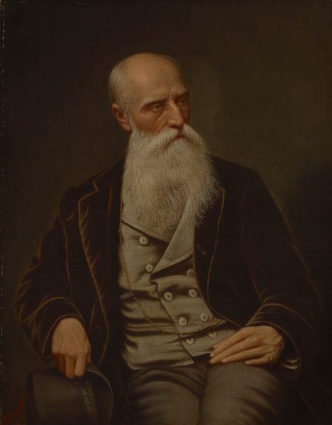 The image depicts a seated man from the waist up. He is facing slightly to his right and looking to his left. The man has a long white beard, closely shaven hair, deeply set eyes, and a slightly furrowed brow. He is wearing a black jacket over a grey waistcoat and trousers. A gold chain is visible around his neck and tucked into the waistcoat. He has a ring on his left hand and is holding a black top hat with his right hand. The background is plain and dark, providing a contrast to his light clothing and beard.