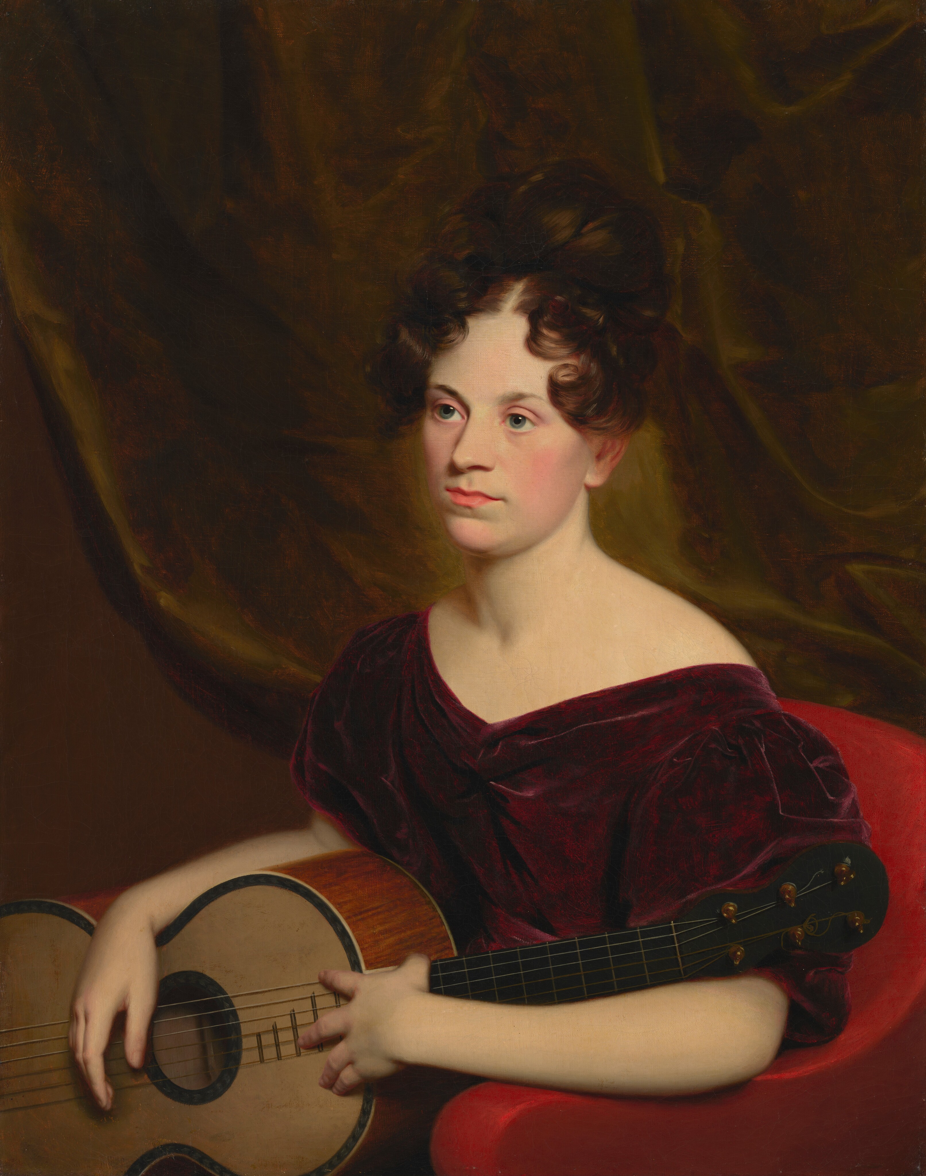 The image depicts the upper body of a woman seated in a red chair against a warm brown backdrop. She is holding a guitar across her lap. Her hair is styled in an elaborate updo with curls and waves framing her face. She has a neutral expression and fair skin with a rosy complexion. She wears a burgundy velvet dress with a wide neckline, revealing her shoulders. The dress's fabric gathers slightly near the neckline. Her hands are resting on the guitar's body and neck, suggesting a relaxed posture. There is no visible jewelry, and the background features a textured drapery.