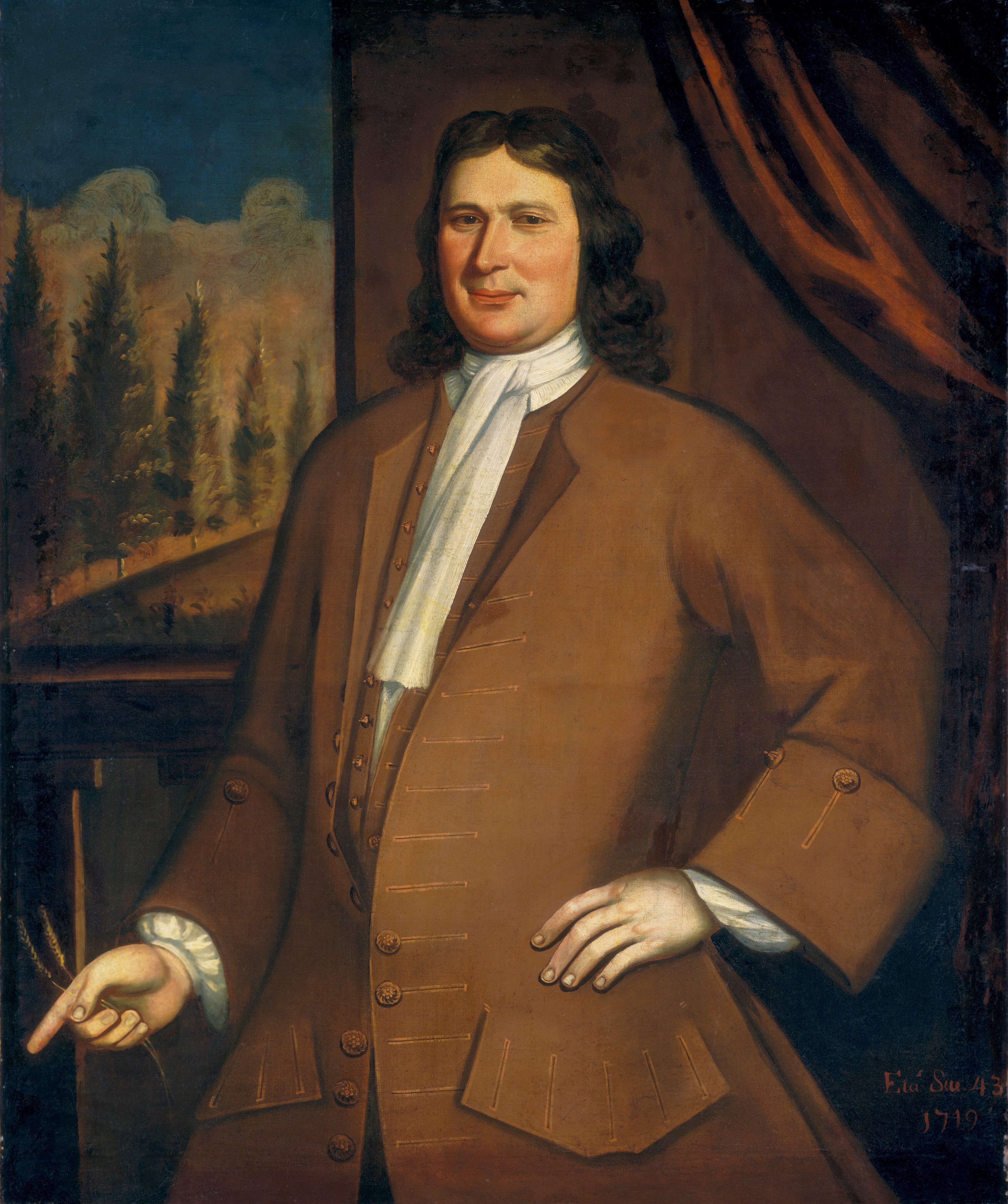This painting depicts a man standing, seen from the waist up. He has one hand resting on his hip and the other pointing downwards towards the bottom-left corner of the painting, holding two small objects that look like pieces of wheat. He has a slight smile, pale skin, a long nose, and long, dark hair falling in waves to his shoulders. He is wearing a brown coat with large buttons and wide cuffs, a matching waistcoat, and a white cravat. The background shows dark red-brown drapery on the right and an outdoor scene on the left, perhaps a window, showing tall, slender trees under a dark blue sky.