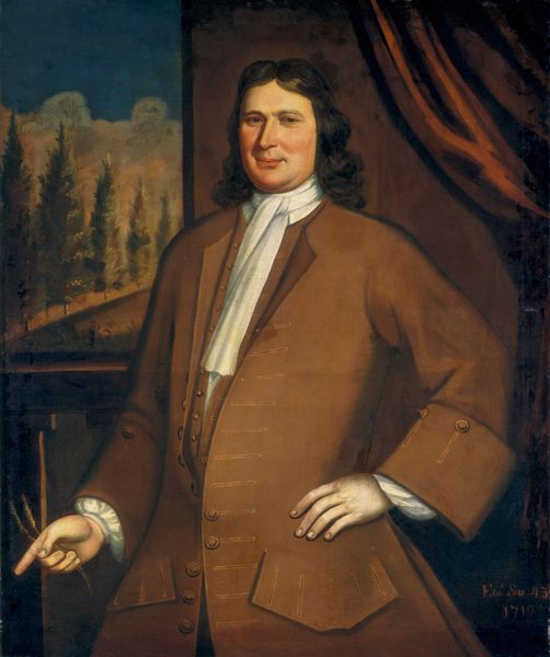 This painting depicts a man standing, seen from the waist up. He has one hand resting on his hip and the other pointing downwards towards the bottom-left corner of the painting, holding two small objects that look like pieces of wheat. He has a slight smile, pale skin, a long nose, and long, dark hair falling in waves to his shoulders. He is wearing a brown coat with large buttons and wide cuffs, a matching waistcoat, and a white cravat. The background shows dark red-brown drapery on the right and an outdoor scene on the left, perhaps a window, showing tall, slender trees under a dark blue sky.