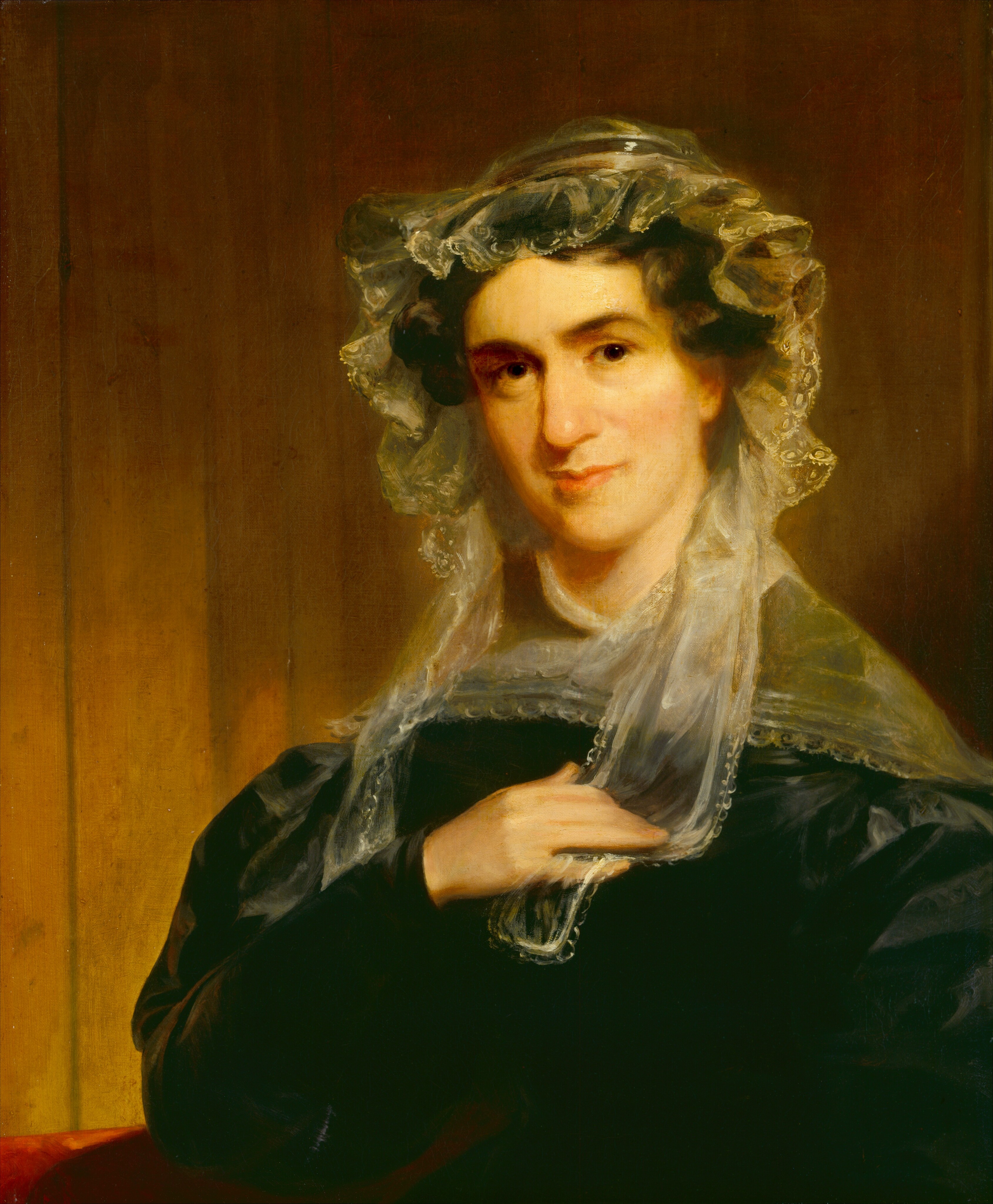 The image shows a bust-length portrait of a seated woman, facing slightly left with her head turned towards the viewer. Her right hand rests on her chest. She has soft facial features, gentle eyes, a straight nose, and slightly pursed lips. She wears a dark garment with long sleeves, complemented by a light-colored lace shawl or collar. Her dark hair is covered by an ornately layered lace cap. The background is a simple, muted brown, likely wood-paneled.