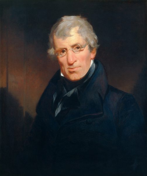 The image shows an older man with a composed expression and grey hair wearing circular spectacles, a dark coat, and a cravat tied around his neck. He is facing slightly to the right and looking towards the viewer. The background is dark and unobtrusive.