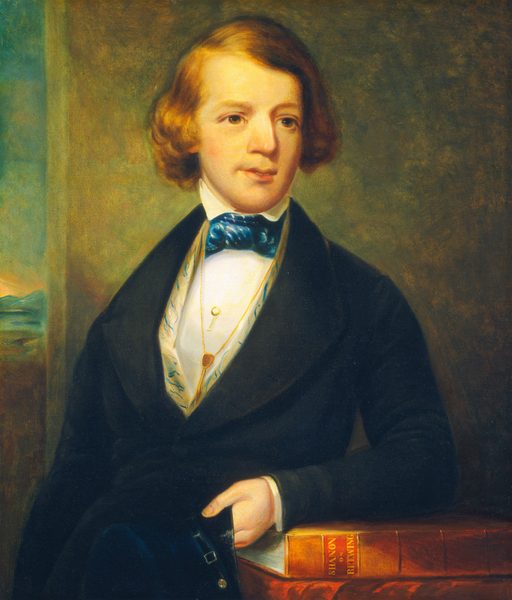 The portrait shows a young man seated with his body turned slightly to the side, facing toward the viewer. He has fair skin and neatly styled, light brown hair that is parted to one side. His face is smooth and youthful, with prominent cheekbones and a defined jawline. He wears a dark coat over a white shirt with a decorative patterned edge. A vivid blue bow tie is neatly tied around his neck. Draped over his waistcoat, a gold chain features a circular pendant and a key-like ornament. His right hand is resting on two large books positioned in front of him, one of which has 'SILVAN' and 'SHONBERG' inscribed on its spine in gold letters. The background consists of a plain, softly shaded wall, with a small portion showing a distant landscape outside a window on the left, depicting a serene natural scene.