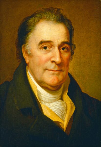 The image shows a man's upper body, focusing on his head and shoulders. He is facing slightly left, looking at the viewer. The man has fair skin, dark eyes, and slightly arched eyebrows. His hair is dark brown with a few grey strands and styled in soft waves. He is wearing a dark jacket, a white high-collared shirt, and a light-colored cravat. The background is a uniform warm tone.