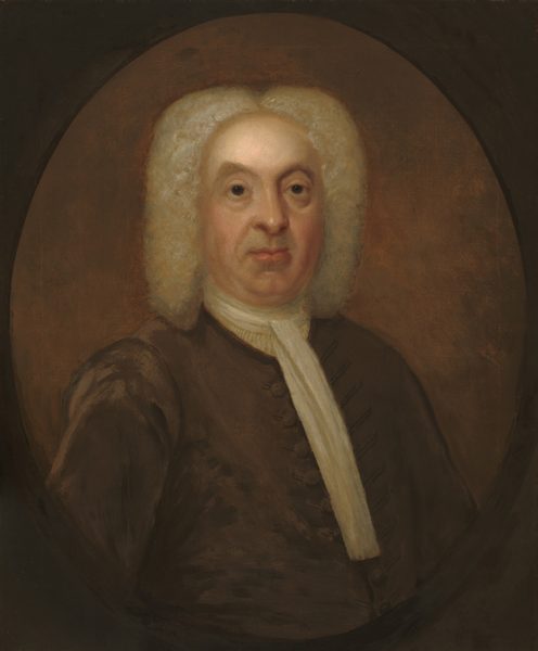 The image features an older man with distinctive facial features and white curly hair, dressed in formal attire against a plain brown background.
