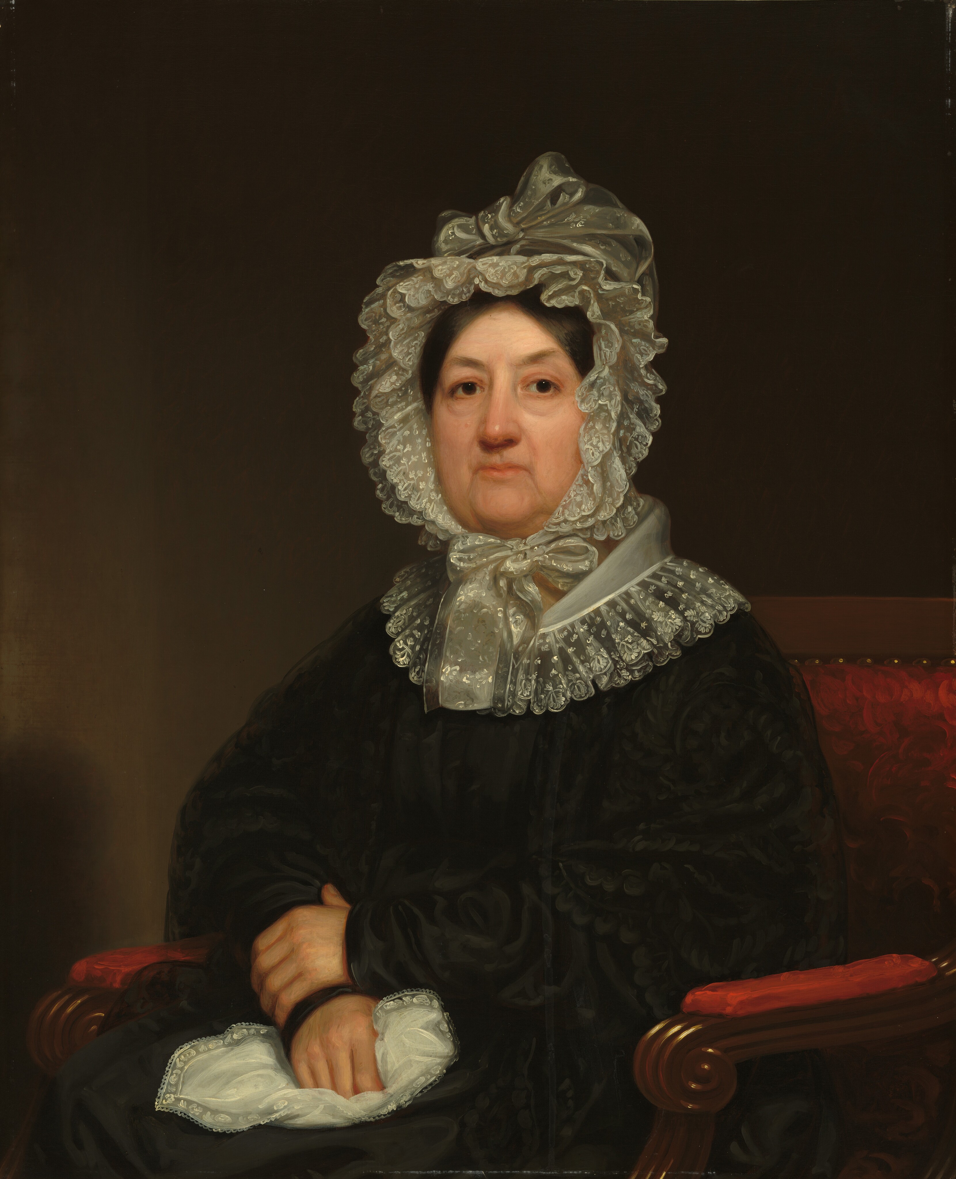 The image shows a woman seated in a red armchair. She is depicted from the waist up with her hands folded in her lap, holding a white lace handkerchief. She has a neutral expression, dark eyes, and wears a dark gown with intricate detailing, a white lace collar, and a lace bonnet with a ribbon. The background is plain and dark.