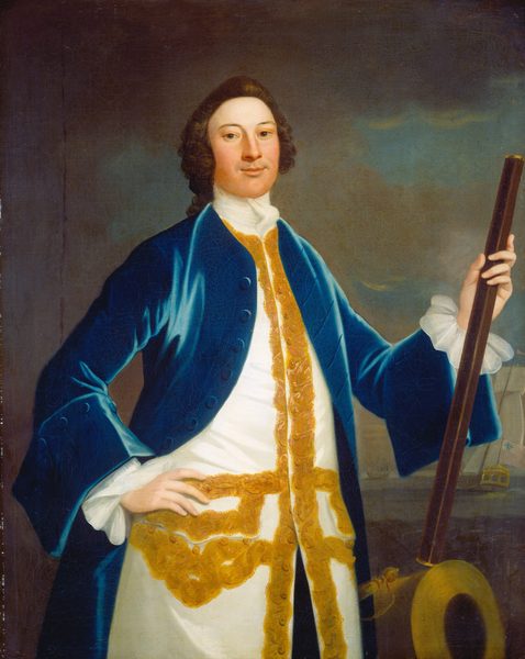 The image shows a portrait of a man from the waist up. He faces slightly left, his left hand on his hip, and his right hand holding a long wooden staff. He has a fair complexion, dark eyes, a rounded face, and dark, curly hair styled in an 18th-century fashion. He wears a blue coat with gold embroidery over a white garment with gold detailing. The coat has wide sleeves with white frilled cuffs and buttons. In the background, there is a hazy seascape with a ship on the right flying a flag, implying a naval context. The backdrop consists of muted blues and browns.