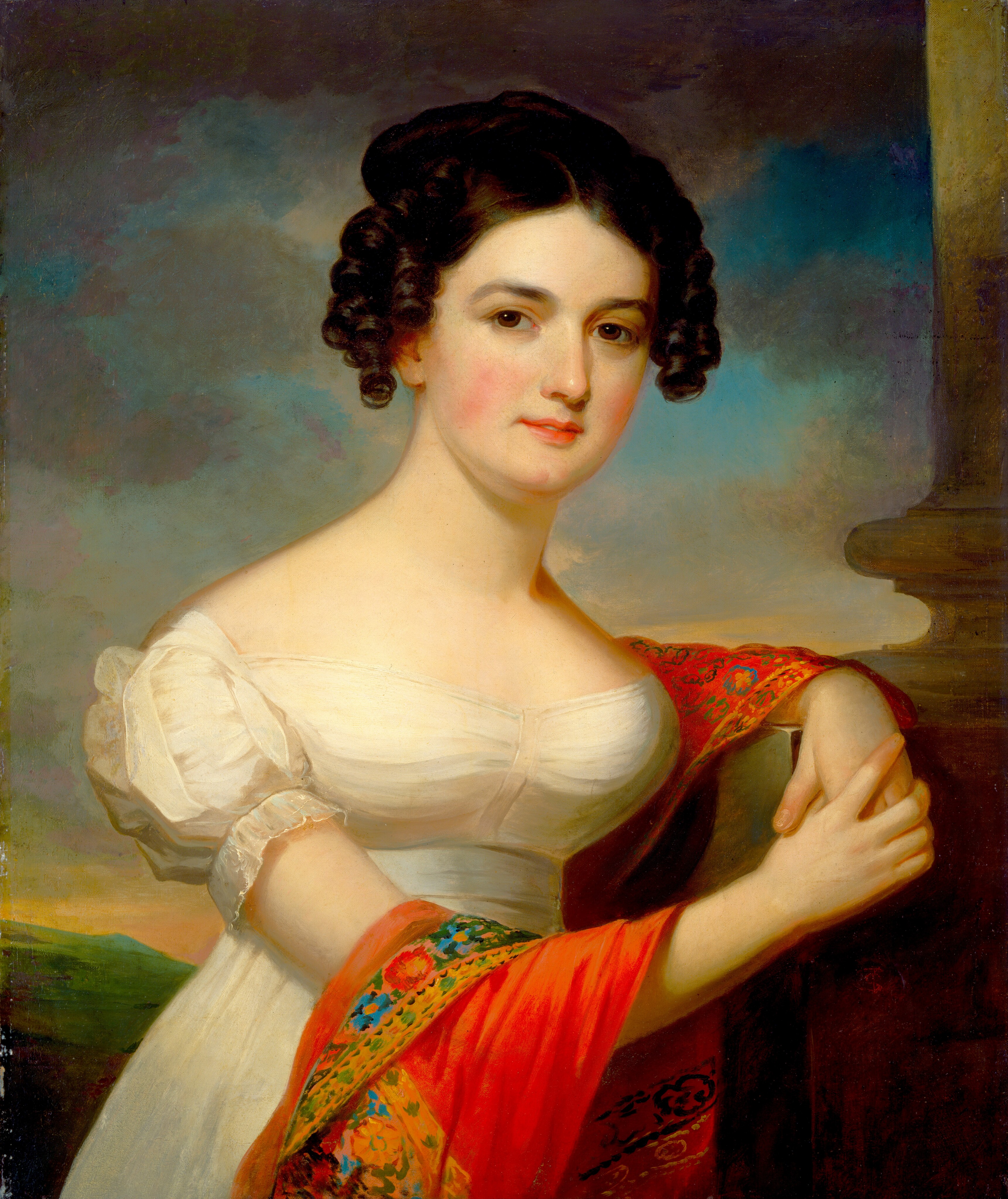 The image shows a woman seated, with her left arm on a stone ledge and her right hand holding her left arm. She has smooth skin, dark eyebrows, rosy cheeks, and dark, curly hair styled around her face and gathered at the back. She wears an off-shoulder white dress with puffed sleeves and holds a vibrant red shawl with a floral pattern. The background shows a dusky sky with soft blue, orange, and pink tones, and some greenery in the distance. A stone column is partially visible on the right side.