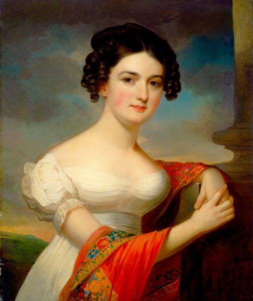 The image shows a woman seated, with her left arm on a stone ledge and her right hand holding her left arm. She has smooth skin, dark eyebrows, rosy cheeks, and dark, curly hair styled around her face and gathered at the back. She wears an off-shoulder white dress with puffed sleeves and holds a vibrant red shawl with a floral pattern. The background shows a dusky sky with soft blue, orange, and pink tones, and some greenery in the distance. A stone column is partially visible on the right side.
