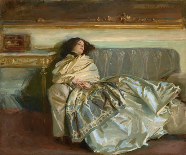 A woman with pale skin and dark brown hair is swathed in silky fabric as she reclines along a gray couch in this horizontal painting. Her head rests along the backrest of the sofa to our left, so her body extends to our right. Her torso is wrapped in a voluminous, ivory-white scarf or wrap. The scarf wraps tightly around her neck, and the bottom edge, near her knees, has a wide, indigo-blue pattern of ovals and vegetal forms. Her shimmering silver gray skirt is painted loosely with baby and sky blue, olive and sage green, and white strokes, and it drapes over the cushions and down the front of the couch. Her hands are clasped so her interwoven fingers rest at her navel. She looks down and off to our right. A table to our left is edged with gold. The wall behind her, above the couch, is painted with long streaks of eggshell white and pale turquoise. The gold frame of a dark painting hanging over the couch spans the width of the composition. Near the upper right corner of the canvas, the artist included a signature and as if he had signed the painting within this painting with loose, dark letters: “John S. Sargent 1911.”