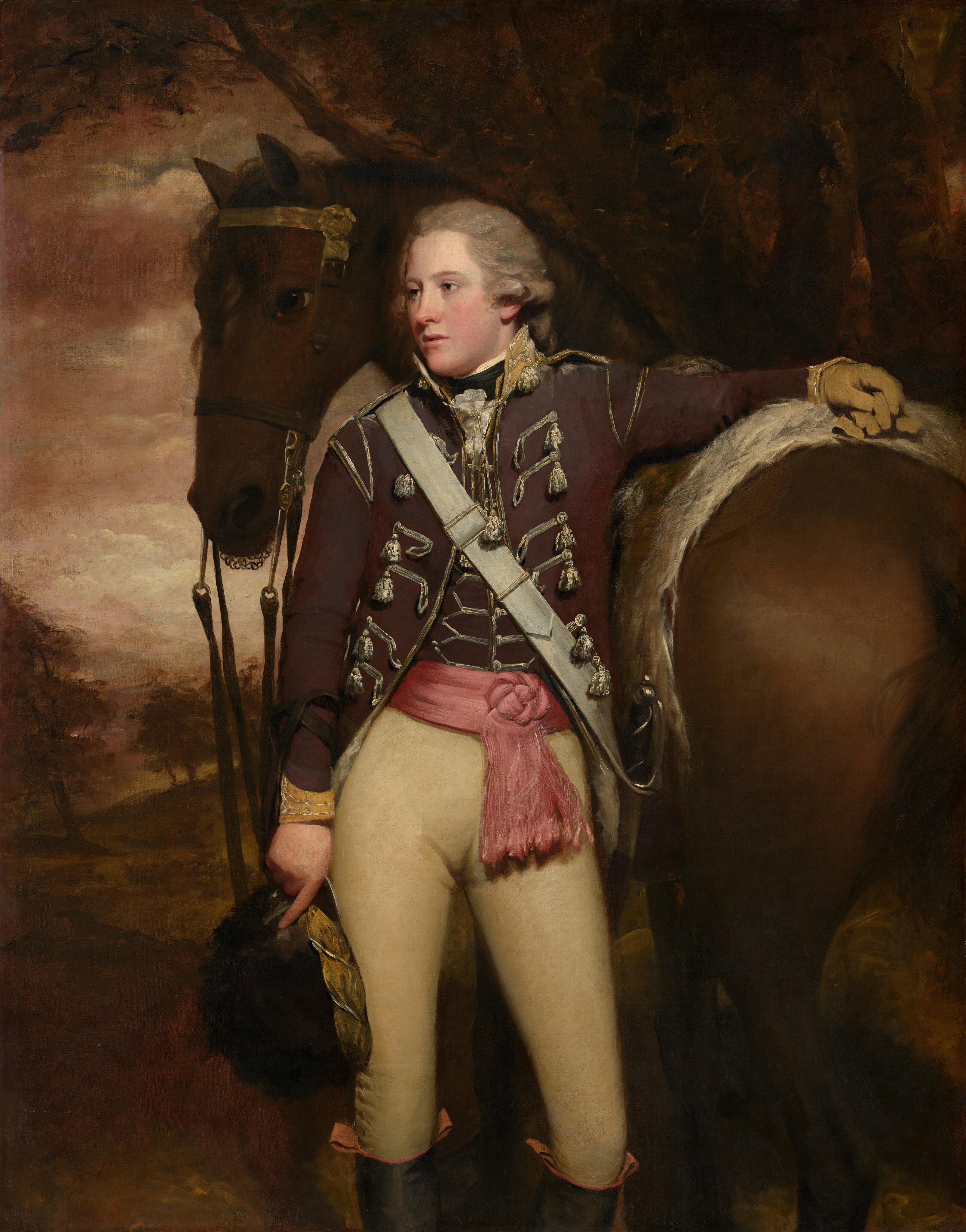 This painting is a portrait of a man standing upright, his chest facing us and his head turned to the left. A horse is positioned directly behind him, and his left arm is lifted, resting on the horse's back. The man has fair skin and wavy gray hair that curls around his ears and seems to be tied behind his neck. He has a slightly flushed complexion, a long nose, pale eyebrows, and lips that seem to be partially open. He wears a formal military uniform, including a brown coat adorned with silver braiding, a brown vest, a pink sash tied at his waist, tan trousers, and tall black boots. He holds a black hat in his right hand. The horse is brown, with its rear visible on the right and its head on the left. The background shows a wooded landscape with dark brown trees and a cloudy red sky.