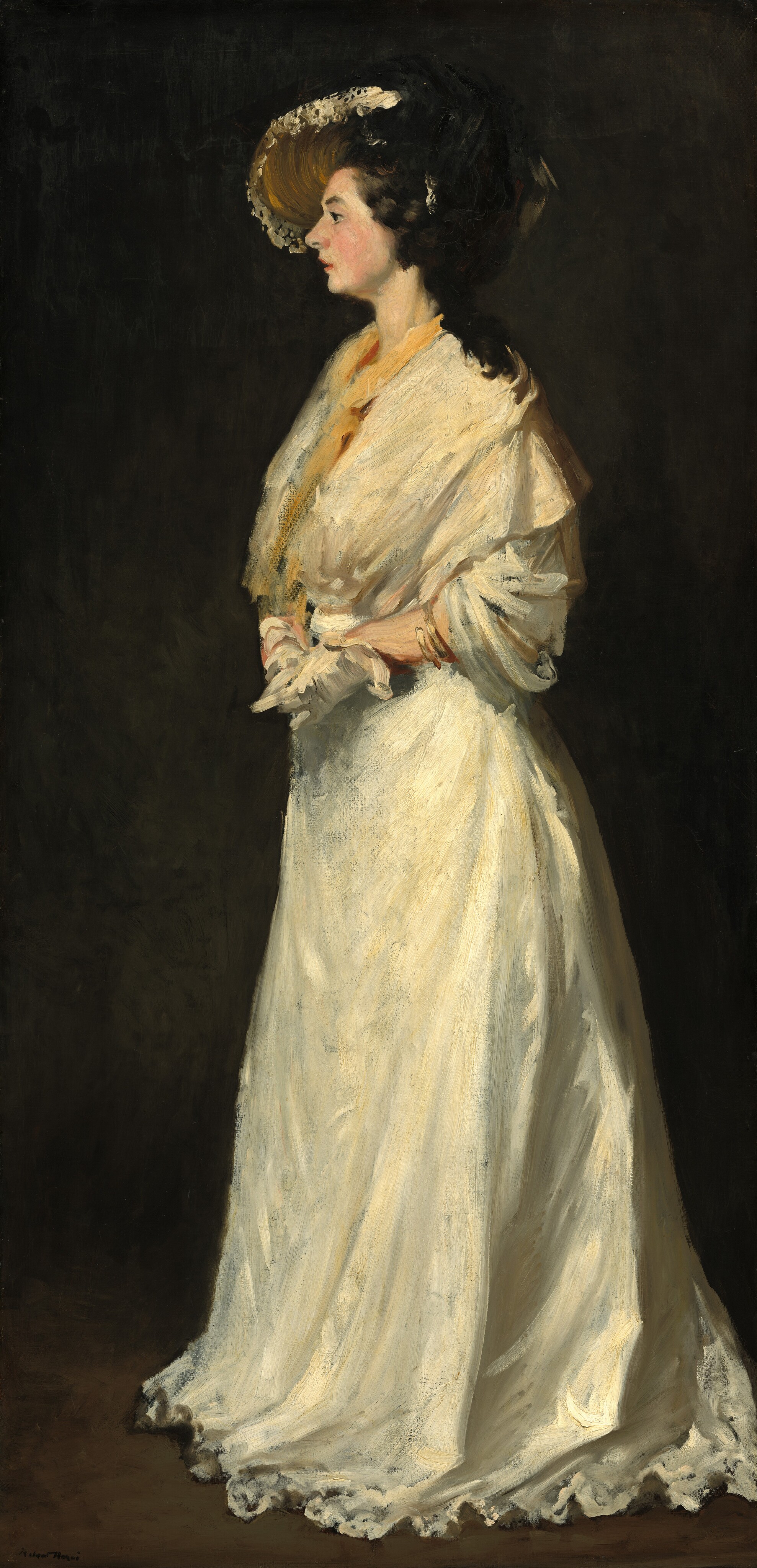 A pale-skinned woman wearing a white gown and hat stands facing our left in profile against a dark background in this vertical painting. The portrait is loosely painted, especially in the clothing and background. The woman’s shoulders are wrapped in a white capelet and golden yellow fabric or lining along the opening. The loose sleeves are gathered near the elbows, and she wears a gold bracelet on the forearm we can see. The skirt falls in heavy folds and pools on the floor in eddying ruffles. Her hands are clasped at her waist, and she wears white gloves. Her black hair is piled atop her head, and locks drape over her shoulder. A wide-brimmed, straw-yellow hat edged with white lace is set at an angle on the far side of her head. She has a strong nose, a slight overbite, parted lips, and rosy cheeks. The background is dark charcoal gray with a band of earthy brown indicating the floor. The artist signed the lower left corner, “Robert Henri.”