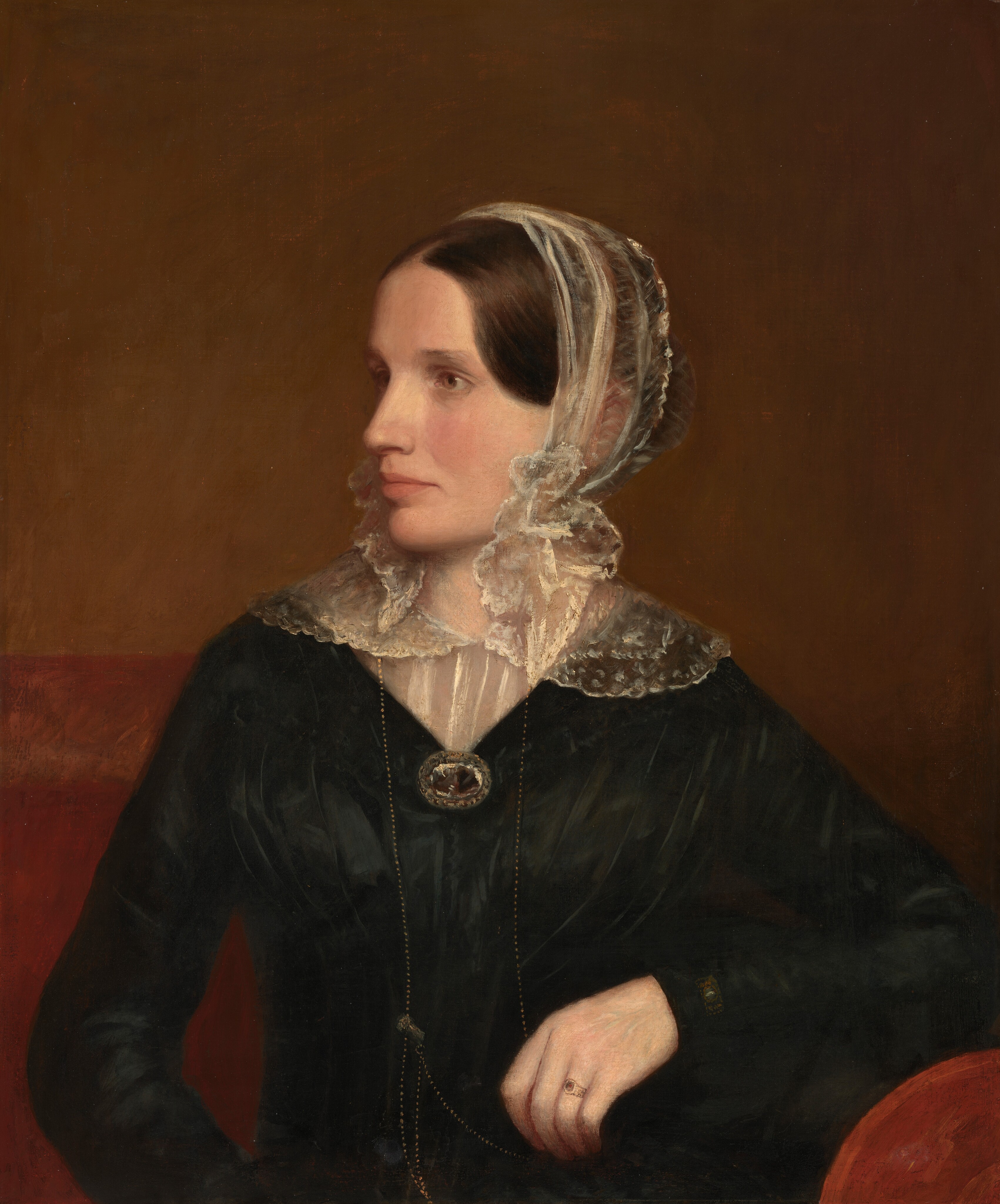 The image shows a seated woman in a side profile. She has soft facial features, brown hair neatly arranged under a lace bonnet, and is dressed in formal attire with a high lace collar. She wears a large oval brooch on her chest and a ring on her left hand. The background is a solid warm brown color.