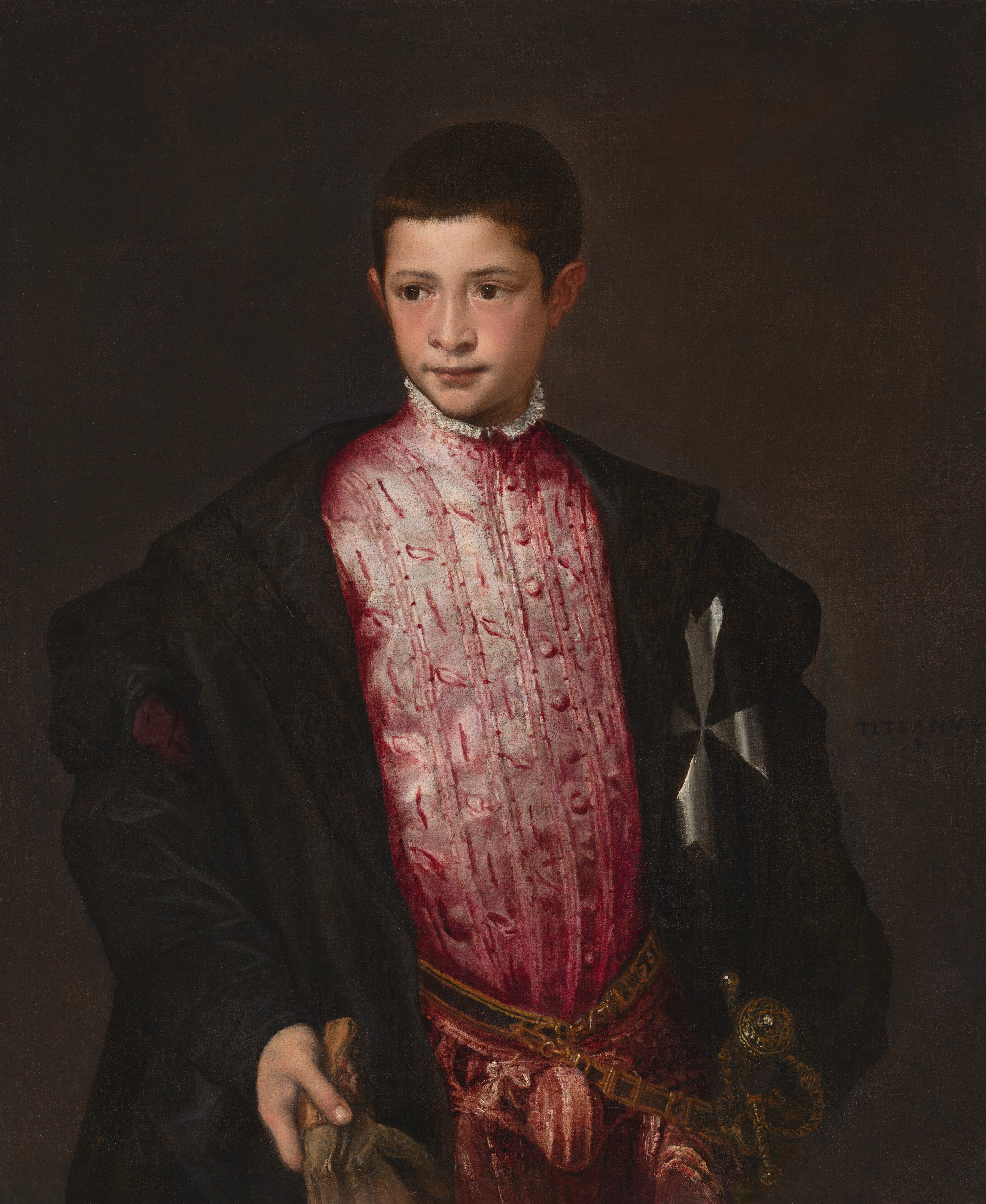 Shown from the hips up, a young boy with pale, peachy skin wears a voluminous, black cloak that nearly falls off his shoulders over a rose-pink tunic, with a sword hanging from one hip in this vertical portrait painting. His body is angled slightly to our right, and he looks off to our left with dark brown eyes under gently arched brows. He has a wide nose, and his pink lips are closed with the corners pulled slightly back. His skin is smooth and his cheeks slightly flushed. His dark brown hair is cut close and comes to a point in front of his ears. His tunic is painted with dusky pink highlights against wine-red to suggest a sheen across a vertically striped, leafy pattern. The garment has a high neck lined with a white ruffle and has a row of buttons down the front. His black coat has wide lapels that reach beyond his shoulders, and the puffy sleeves gather on his arms. The coat has a silvery-white cross over the chest to our right. The left and right arms of the cross are lost in the folds but the arms at the top and bottom are forked at the ends. The boy’s pine-green belt is edged with gold, and the hilt of the sword is angled toward us on his left hip, to our right. He holds one fawn-brown glove in his right hand, to our left, and his other arm disappears behind the folds of his coat. The background is deep olive green, almost brown. The artist signed the painting with dark paint near the right edge of the canvas near the boy’s shoulder, “TITANVS F.”