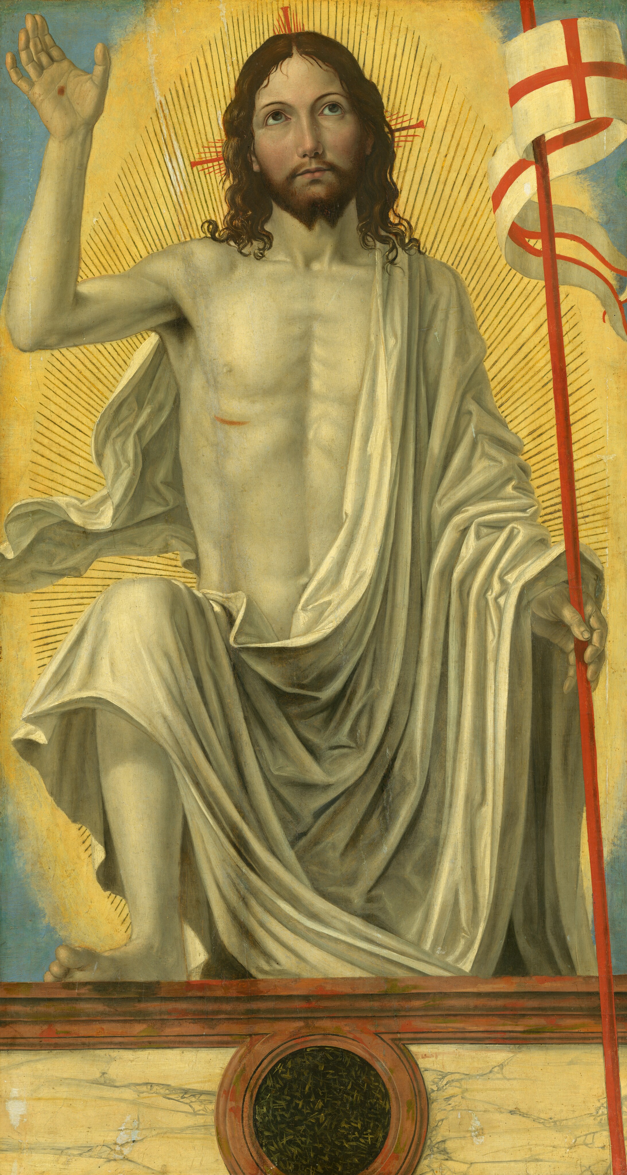 A man draped in a white cloth steps toward us onto a ledge as he holds a red staff with a white banner in one hand and holds up the other hand to show a wound on the palm in this vertical painting. His pale face has a yellow cast but he has rosy cheeks, and his body is eggshell white. Near the upper left corner of the painting, his raised right hand, to our left, is held palm out to show it has been pierced. The cardinal-red staff he supports in his other hand has a long, fluttering banner with a red cross against a white background. He bends his right knee deeply, so his bare foot rests on a low wall or side of a casket in front of him. He looks up with brown eyes under curved eyebrows. He has a straight nose, and his small, rose-pink mouth is closed. He has shoulder-length, brown curly hair and a forked beard. His voluminous ivory-white cloth drapes over one shoulder and hangs in deep folds across the arm holding the banner and the opposite knee. He has a bloody gash over his right ribs. His entire body is surrounded by radiating lines encompassed in a glowing, golden halo. The corners around the halo are celestial blue. The ledge where he props his foot is painted to look like yellow marble veined with gray, framed with fawn-brown molding along the top edge. A round, black medallion flecked with celery green at the front center of the panel is also surrounded with with brown molding.