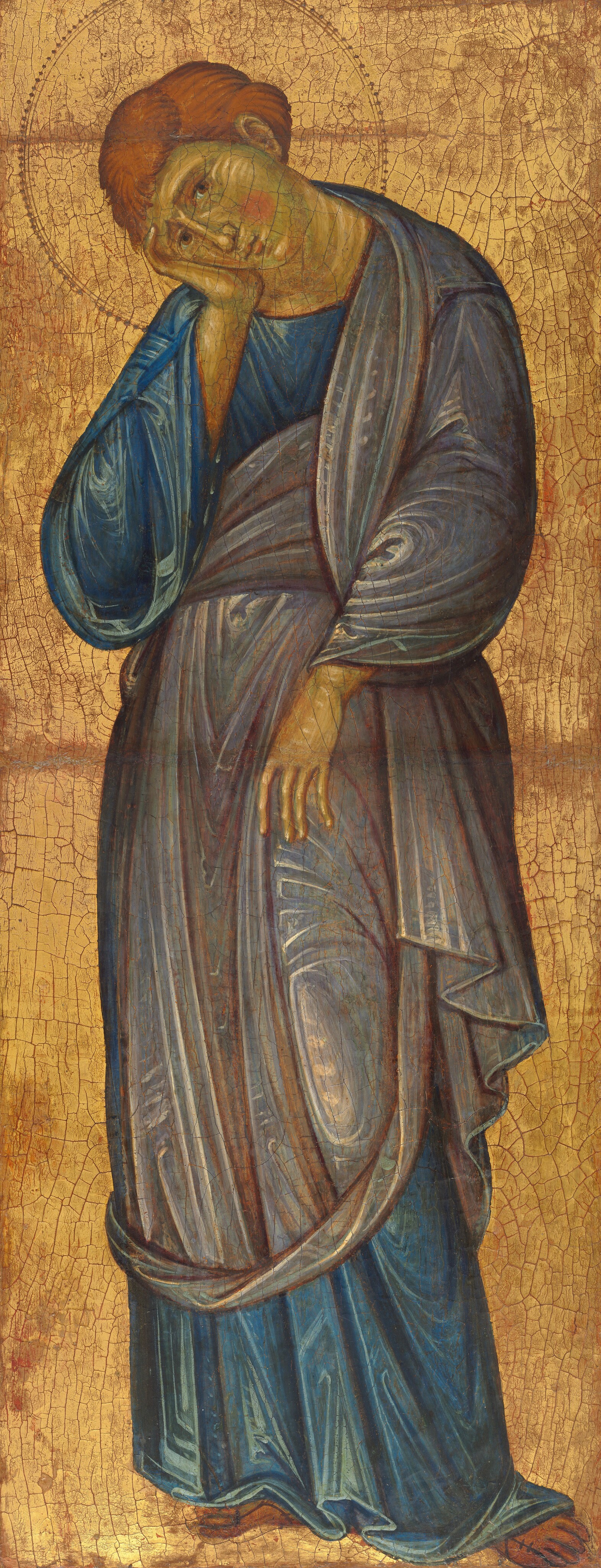 Shown against a shiny gold background, a cleanshaven young man with carrot-orange hair stands with his head tipped sharply to rest in one hand in this vertical painting. His skin is pale with a noticeably green tint. He tips his head to our left and looks at us with brown eyes. His left cheek flushes with a bright pink circle, and four vertical white strokes indicate creases in the flesh above the cheekbone. Visible strokes in butter yellow pick out his chin, nose, wrinkles on his furrowed forehead, and the fingertip of his pinky finger, along his cheek. His long-sleeved, lapis-blue garment falls in stylized folds to his bare feet. The lighter, slate-blue mantle wraps around his torso to his knees. The long fingers of his left hand, on our right, rests against his thigh. White highlights create swirling, angular patterns in the folds of the garments. A halo around his head is punched and incised into the gold background. A network of cracks runs across the gold background, and the red layer underneath is visible in some areas.