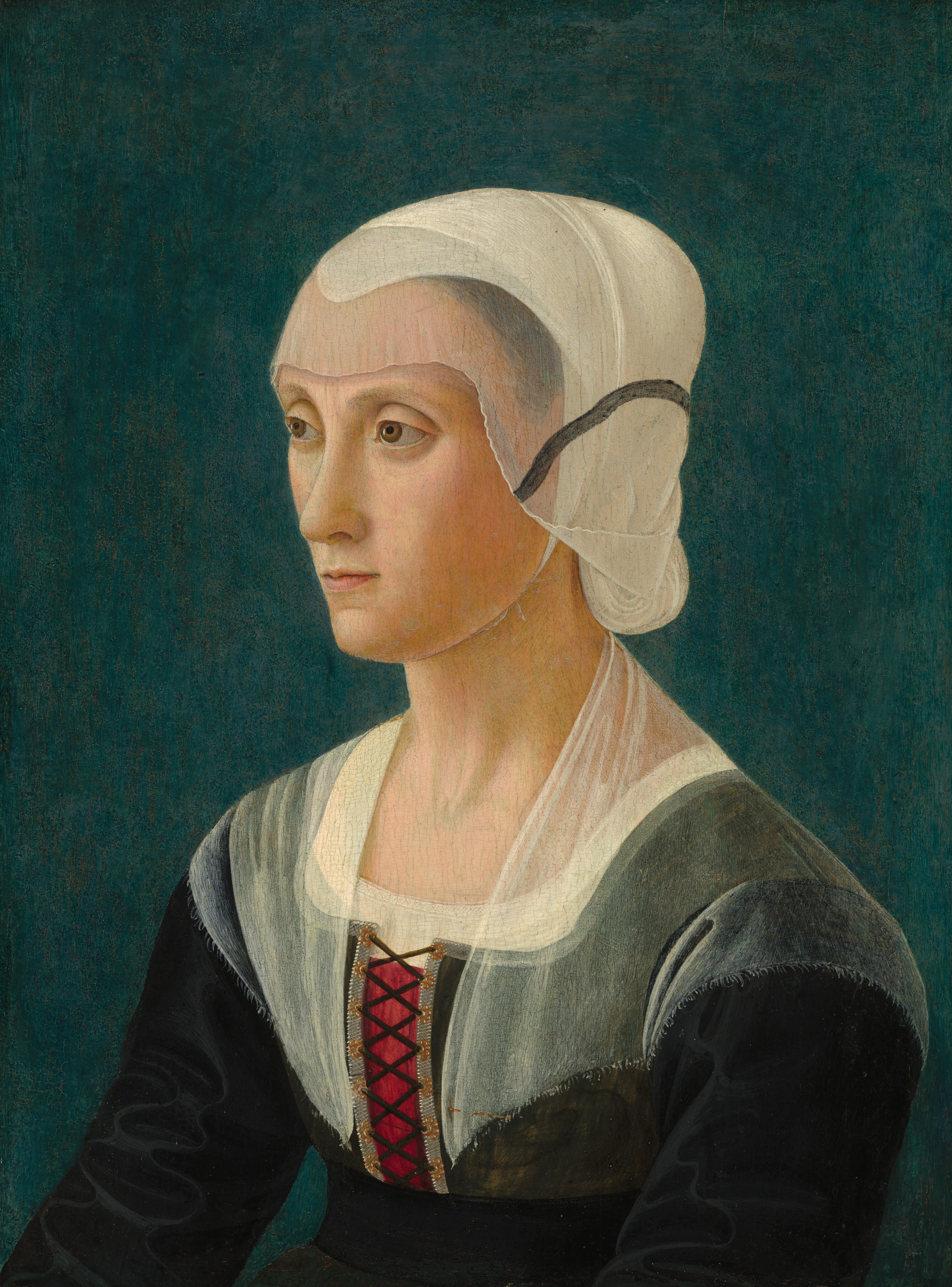 Shown from the chest up against a dark, teal-blue background, a light-skinned woman looks off to our left left in this vertical portrait painting. Her arms extend slightly in front of her, as if they rest on her knee. Her face turns almost in profile to our left as she gazes in that direction with hooded light brown eyes under curved brows. She has a sloping nose, a pointed chin, and her thin, light pink lips are closed. A white covering over her hair, which has been pulled back and up, is layered under a translucent ivory-white veil that covers her forehead and the sides of her face. A charcoal-gray stripe on the veil creates a line from her cheek to the back of her head. Her dress has a black bodice and sleeves, and the chest and the tops of her shoulders are covered with another veil. A patch of burgundy red shows through where the dress is laced up over the chest.
