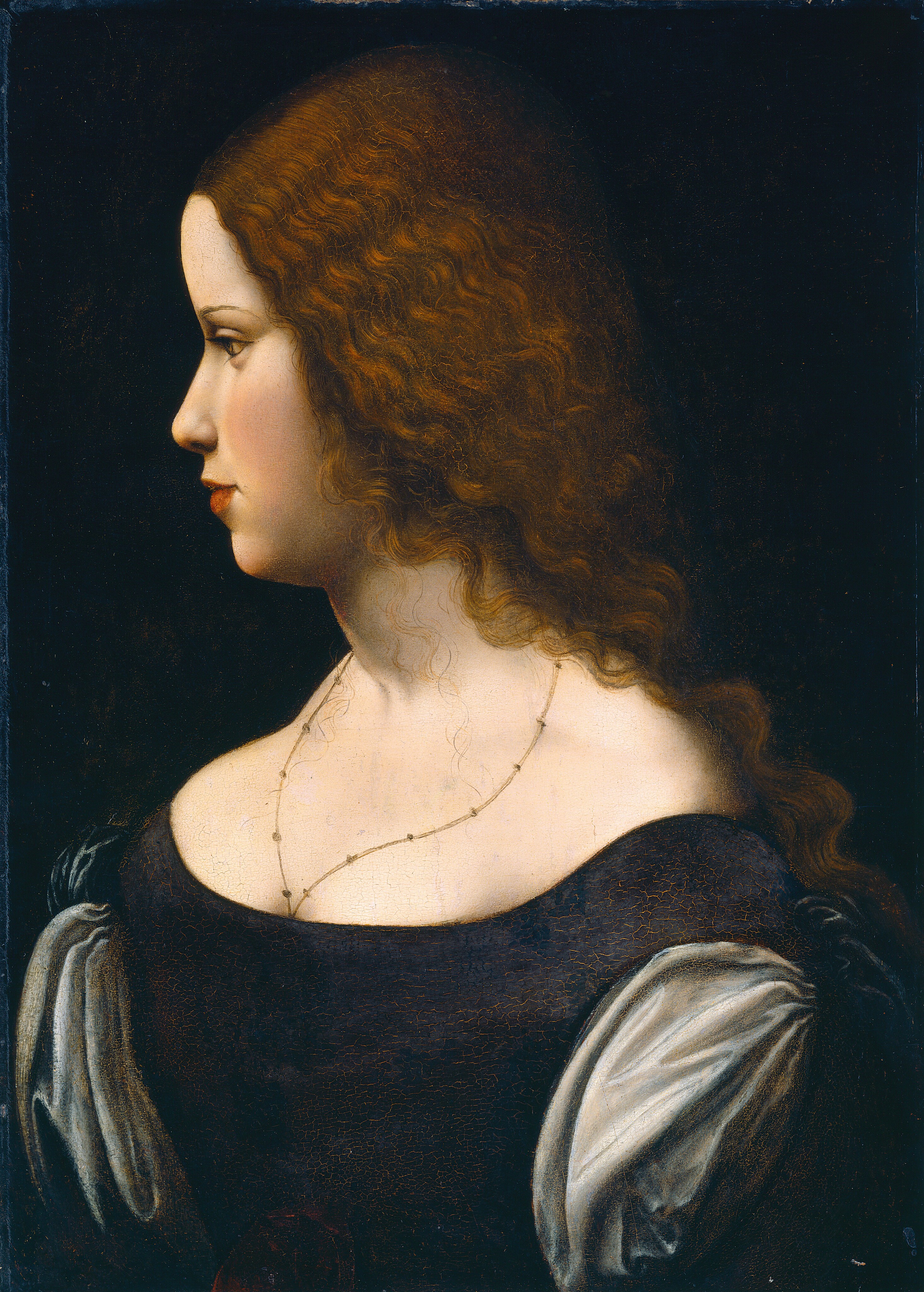 Shown against a black background, the head and shoulders of a young lady with pale, peachy skin and long, wavy brown hair fill this vertical portrait painting. The woman faces our left in profile and she looks off in that direction with light brown eyes, her head tipped slightly forward. Her skin is smooth and her full cheeks are lightly flushed. The eyebrow we can see is thin and arched. She has a small, straight, rounded nose and her full terracotta-red lips are closed. Her hair is gathered back over her shoulders, but a few fine, curling tendrils fall over her chest. She wears a thin necklace that disappears into the cleavage of her low-cut, navy-blue bodice, which has sky-blue, puffed sleeves. The back of her head is lost in shadow against the dark background.