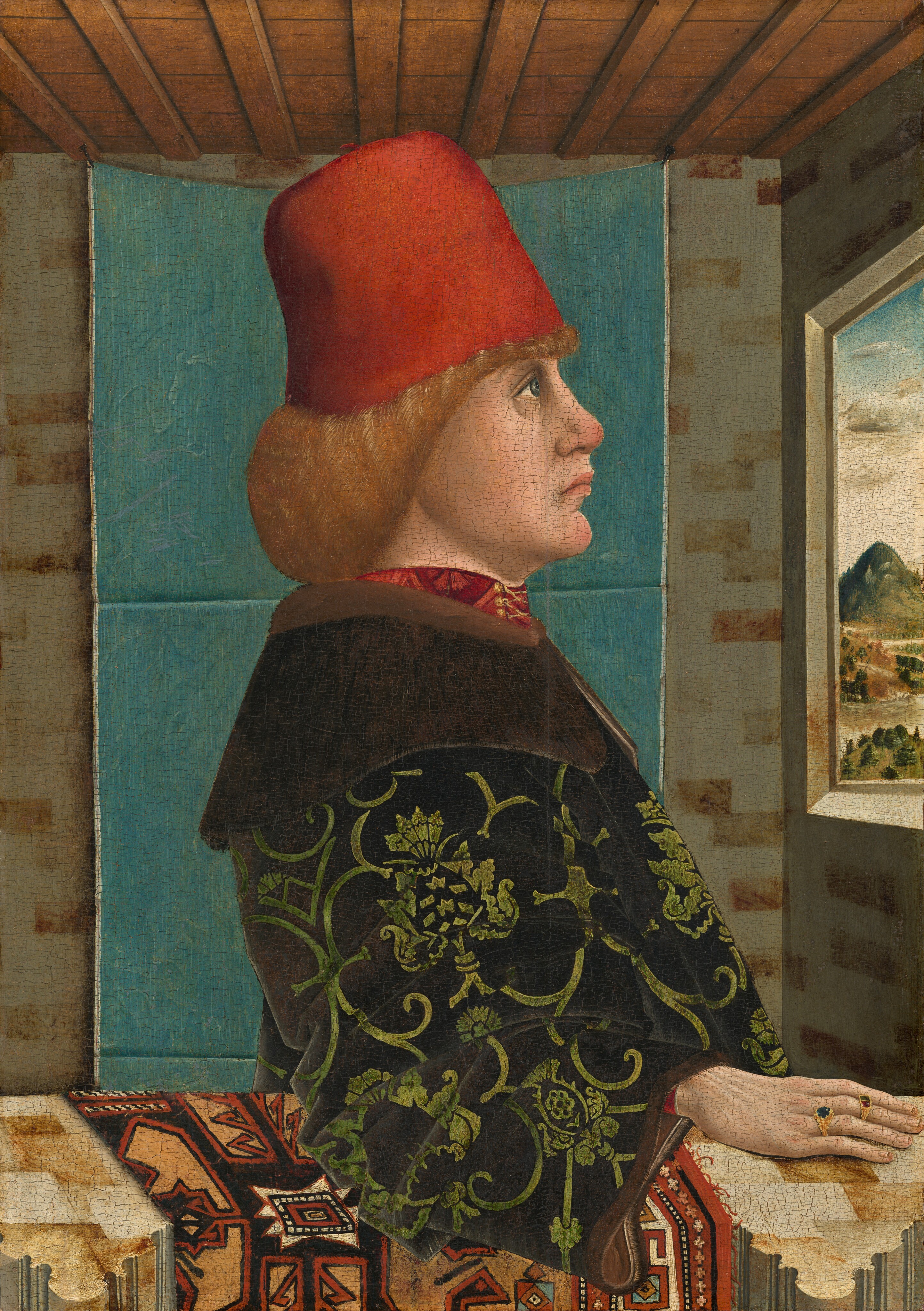 This painting shows a man from the waist up, standing in the corner of a room. He faces a window on the right that displays an outdoor landscape with green mountains and trees under a blue-and-white sky. The man has a pale complexion, with a large nose, prominent chin, deep-set eyes, and pink lips set in a slight frown. He has reddish-blond shoulder-length hair under a vivid red hat. He is dressed in a dark robe with green floral patterns over a red garment that is visible around his neck. The man's right hand rests on a surface next to him, and he wears two visible rings, one with a blue stone and one with a red stone. The background includes a brick-patterned wall in shades of cream and tan, a blue curtain or tapestry, and wooden ceiling beams.