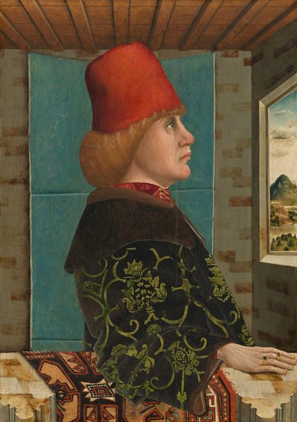This painting shows a man from the waist up, standing in the corner of a room. He faces a window on the right that displays an outdoor landscape with green mountains and trees under a blue-and-white sky. The man has a pale complexion, with a large nose, prominent chin, deep-set eyes, and pink lips set in a slight frown. He has reddish-blond shoulder-length hair under a vivid red hat. He is dressed in a dark robe with green floral patterns over a red garment that is visible around his neck. The man's right hand rests on a surface next to him, and he wears two visible rings, one with a blue stone and one with a red stone. The background includes a brick-patterned wall in shades of cream and tan, a blue curtain or tapestry, and wooden ceiling beams.