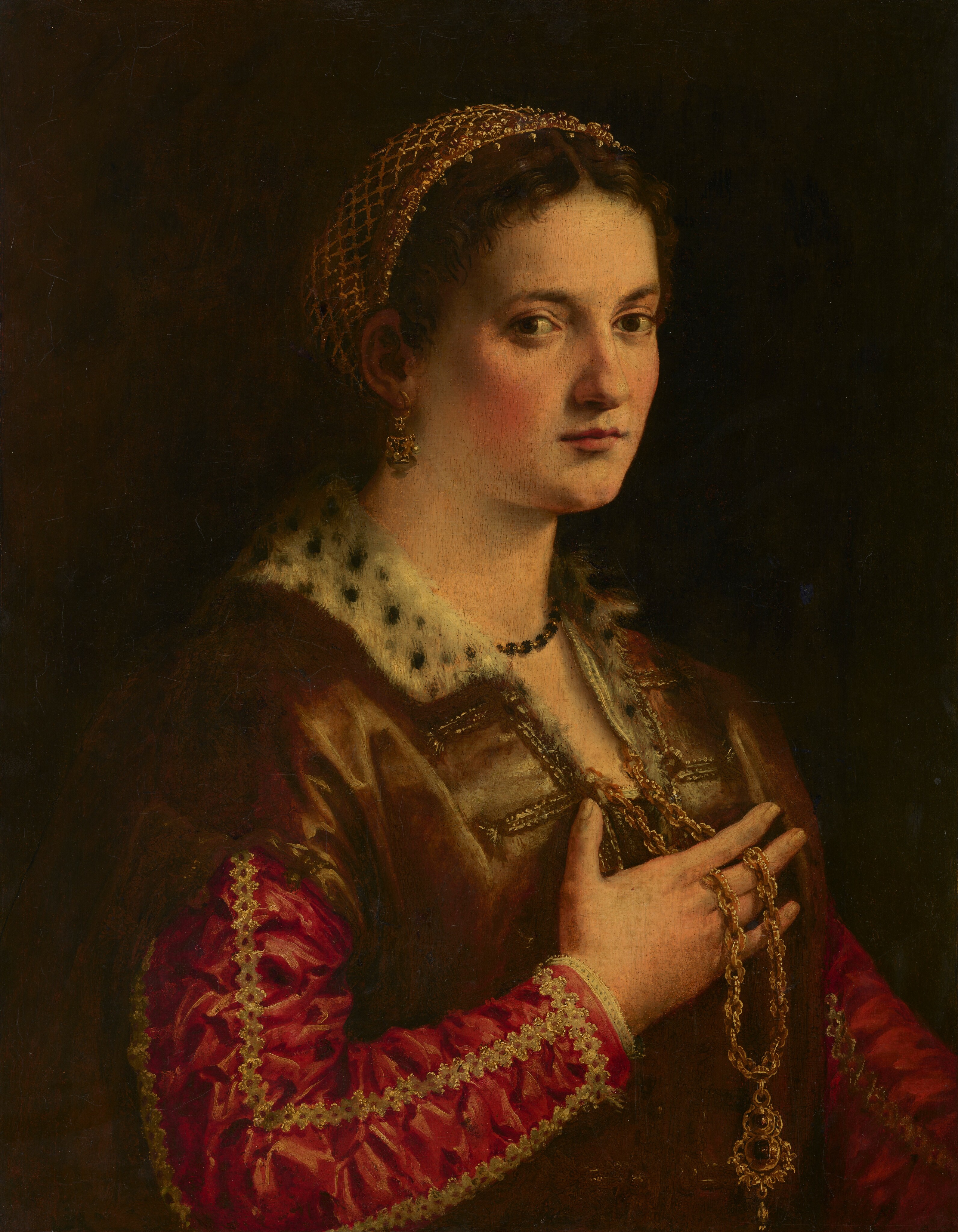 Shown from the waist up, a young woman with pale, faintly yellow-tinged skin and flushed cheeks looks at us in this vertical portrait painting. The woman’s body is angled to our right, and she gazes out from the corners of her brown eyes. She has a long nose, and her pale red lips are closed. Short strands of her dark brown hair curl onto her forehead, and the rest of her hair is covered by a net-like gold headdress fringed with pearls. More pearls dangle from a gold earring hanging from the ear we can see. A necklace with small black gems encircles her neck. Her brown garment is lined with black-dotted white fur at the neck and down the open front. Under it, her raspberry-pink, long-sleeved dress is lined with gold trim. A long, thick gold chain lies between the fingers of her right hand, closer to us, and a gold and jewel medallion dangles from the bottom. he is lit from our right against a dark brown background.