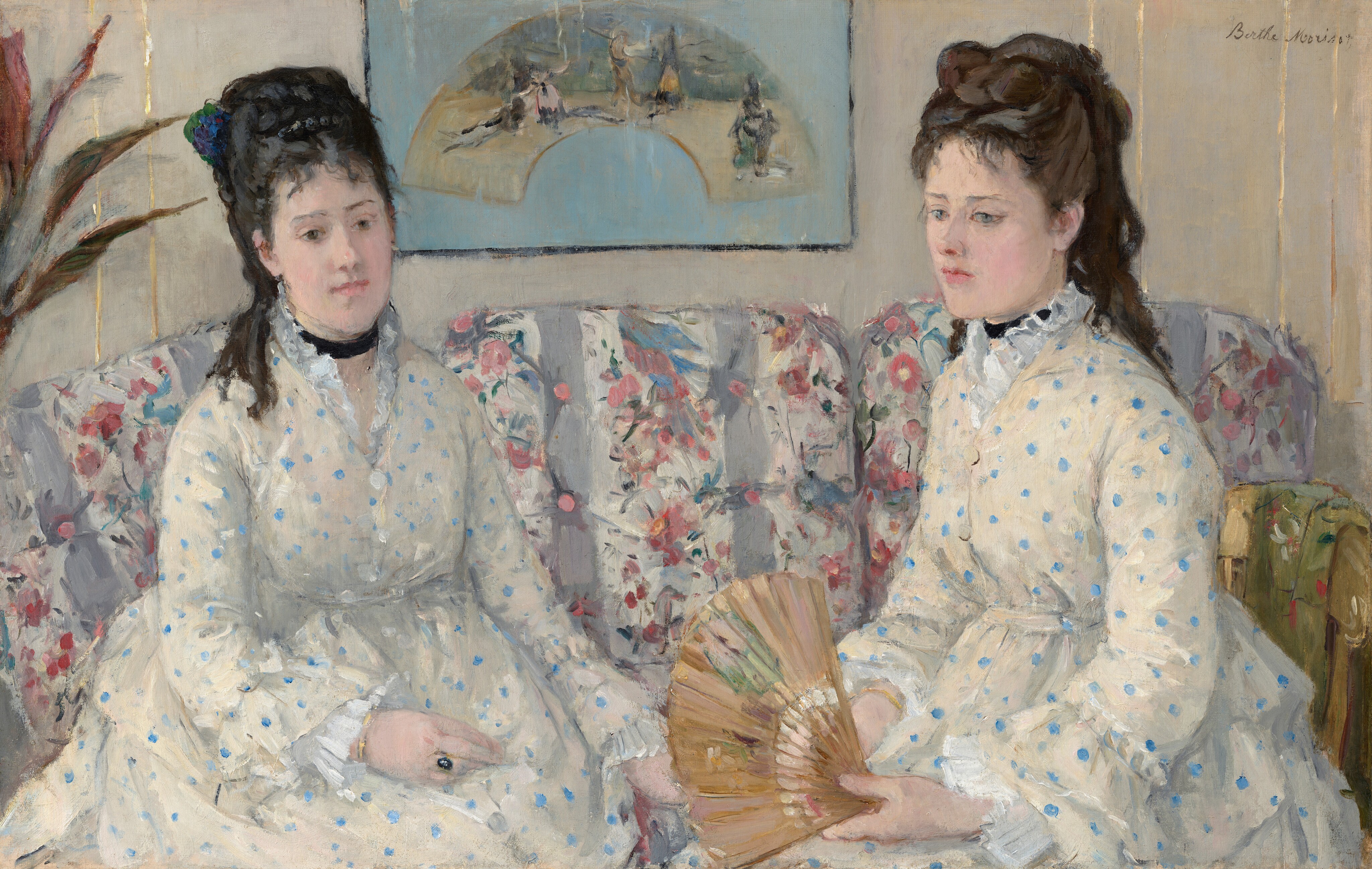 Shown from the lap up, two women with pale skin and dark hair pulled up and back, wearing white dresses with baby-blue polka dots, sit on a couch in this horizontal portrait painting. Both women have straight, dark brows, delicate noses, smooth skin, and their small, rose-pink mouths are closed. Wispy bangs brush their foreheads, and their hair is piled high with long ringlets falling down their backs. Both wear black ribbons like chokers around their necks. Their dresses have high necks with ruffles along the necklines, ruffles at the wrists, and are gathered under the bust. They sit angled in toward each other. The woman on our left has black hair, and she looks down and to our right with dark eyes. She wears a gold ring with a dark, oval stone on one hand resting in her lap. The other woman has chestnut-brown hair and looks down and off to our left with ice-blue eyes. She holds an open fan in her hands in her lap. The fabric on the couch has vertical white and lilac-purple stripes, and is overlaid with a pattern of pink flowers and green leaves. A framed picture hanging on the bone-white wall behind the women shows an arched painting against a sky-blue background. A few spiky leaves from a houseplant are cut off by the left edge of the painting. The portrait is loosely painted throughout, especially in the couch and background. The artist signed the painting in the upper right corner, “Berthe Morisot.”