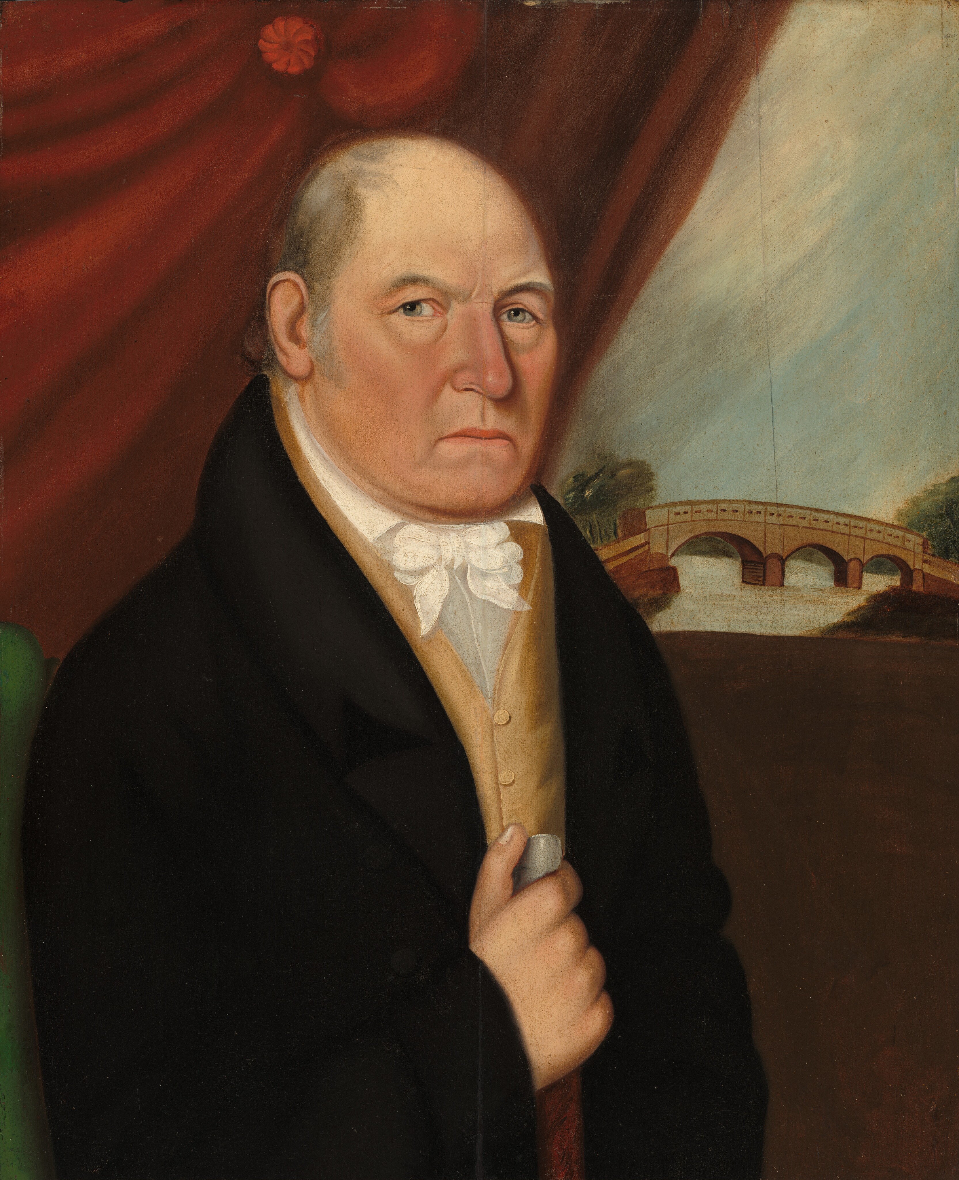 The image shows a man facing forward with his body slightly angled to the right. He has an expressionless face with a slightly furrowed brow, a pronounced nose, thin lips, and light eyes. His hair is balding on top with some hair around the sides and back. He is dressed in formal attire, including a black coat, a light brown vest, a white shirt with a high collar and a white cravat. He holds a cane or walking stick with his right hand. He is seated near a green surface, possibly a chair or bench. The background features a painted backdrop of a pastoral scene with a wooden bridge over a river and trees in the distance. A red draped curtain runs across the top of the image.