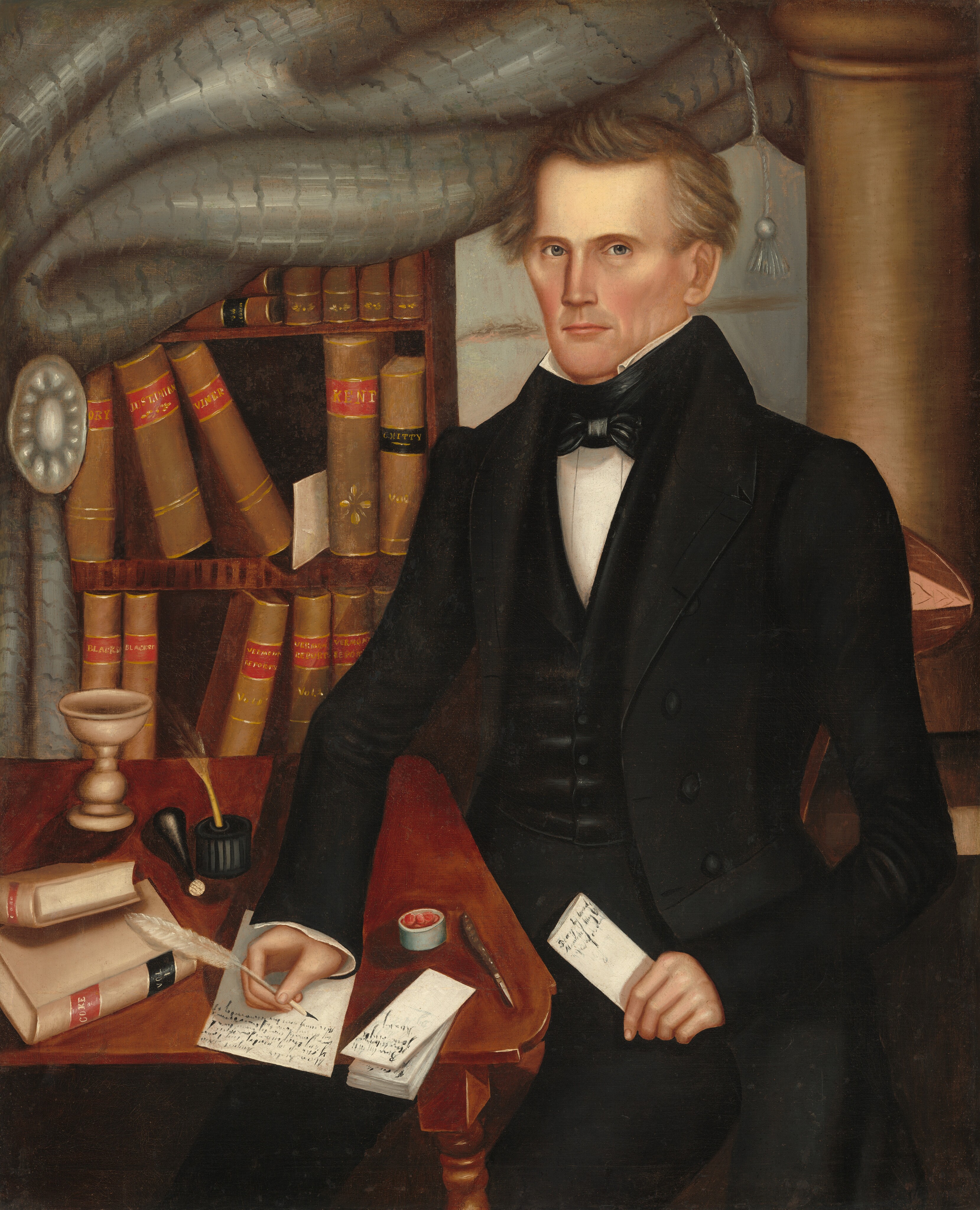 The image shows a man seated at a wooden desk with a formal posture. He has sharp facial features and light brown hair styled with waves. He wears a black coat, white shirt, black waistcoat, and a black bow tie. On the desk are sealed books, a quill pen in an inkwell, and a folded paper. The man holds a piece of paper in his left hand. Behind him is a bookshelf filled with books and documents, partially covered by a large curtain. An architectural column is visible in the background.
