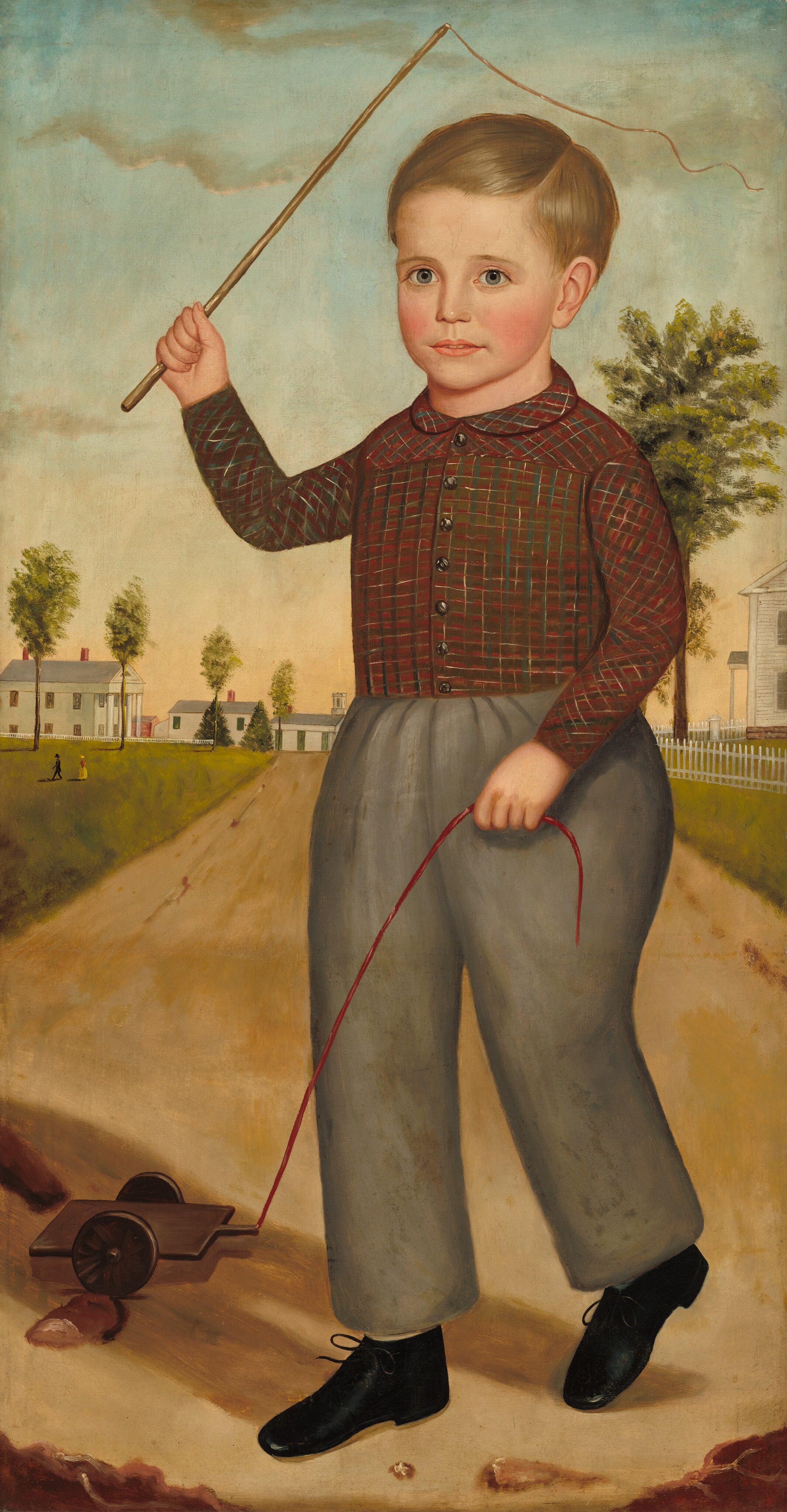 The image shows a young boy standing on a dirt path, holding strings attached to a wooden toy with wheels. He is raising his left hand with a stick. The boy has short, neat light brown hair, fair skin, rosy cheeks, and bright eyes. He wears a red and green checkered shirt, gray trousers, and black shoes. In the background, there are white houses with red chimneys, trees, a sky with clouds, and a picket fence.
