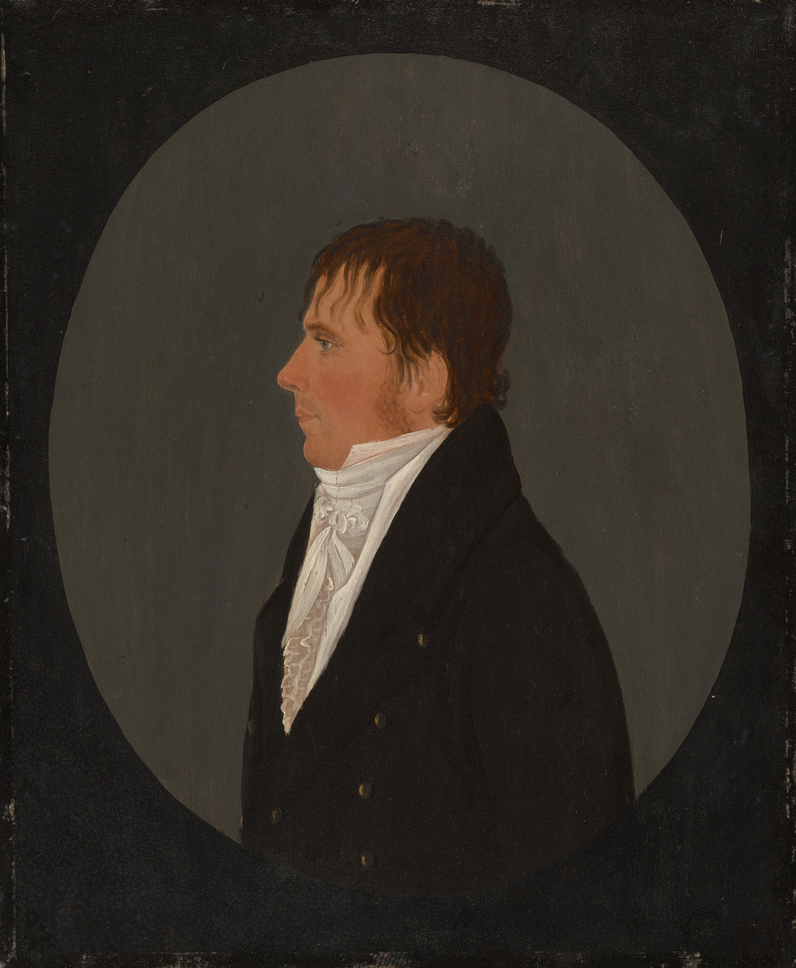 The image shows a portrait of a man from the chest up, in a side profile position facing left. He has a fair complexion with distinct facial features, including a straight nose and a strong jawline. His hair is brown and styled casually. He is wearing a black coat over a white high-collared shirt with ruffles. The background is a solid, muted gray tone.