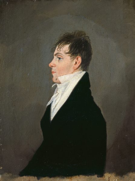 The image is a profile view portrait of an individual, showing the upper half of the body. The person is positioned sideways, facing left. He has a fair complexion, with a prominent nose and eyes that are directed straight ahead. His hair is brown, slightly tousled, and somewhat curly at the ends. The person is wearing a dark-colored jacket with a high-collared, white shirt beneath it. There are no visible pieces of jewelry or objects being held. The background is a muted, monochrome shade.