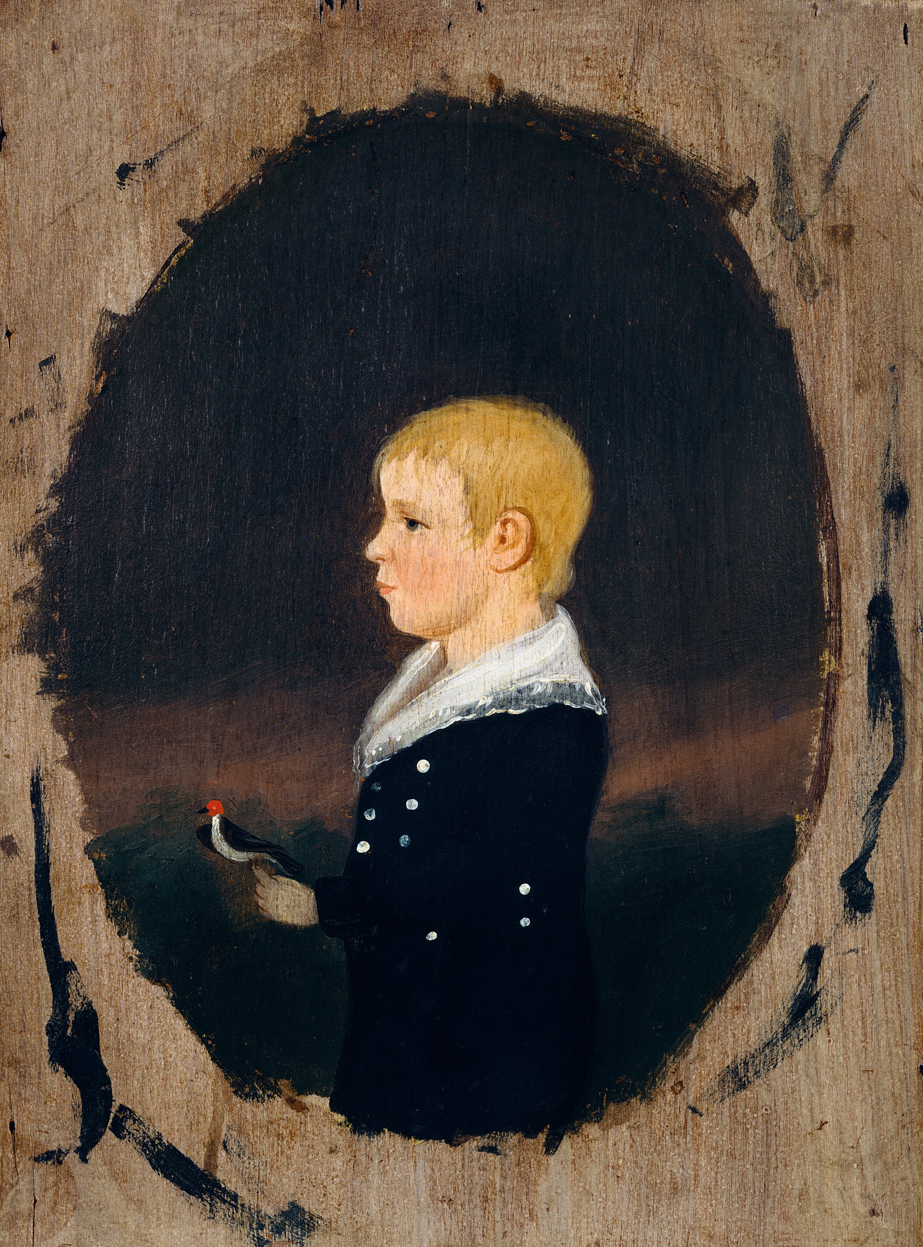 The image is a portrait showing the upper half of a young boy's body, depicted in profile. He is holding a small bird with a red head in his right hand. The boy has short, light blonde hair and features a delicate profile with a slightly upward-tilted nose. He is wearing a dark-colored outfit adorned with small white buttons and a wide white collar with intricate details. The background is a dark oval shape surrounded by a wooden texture.