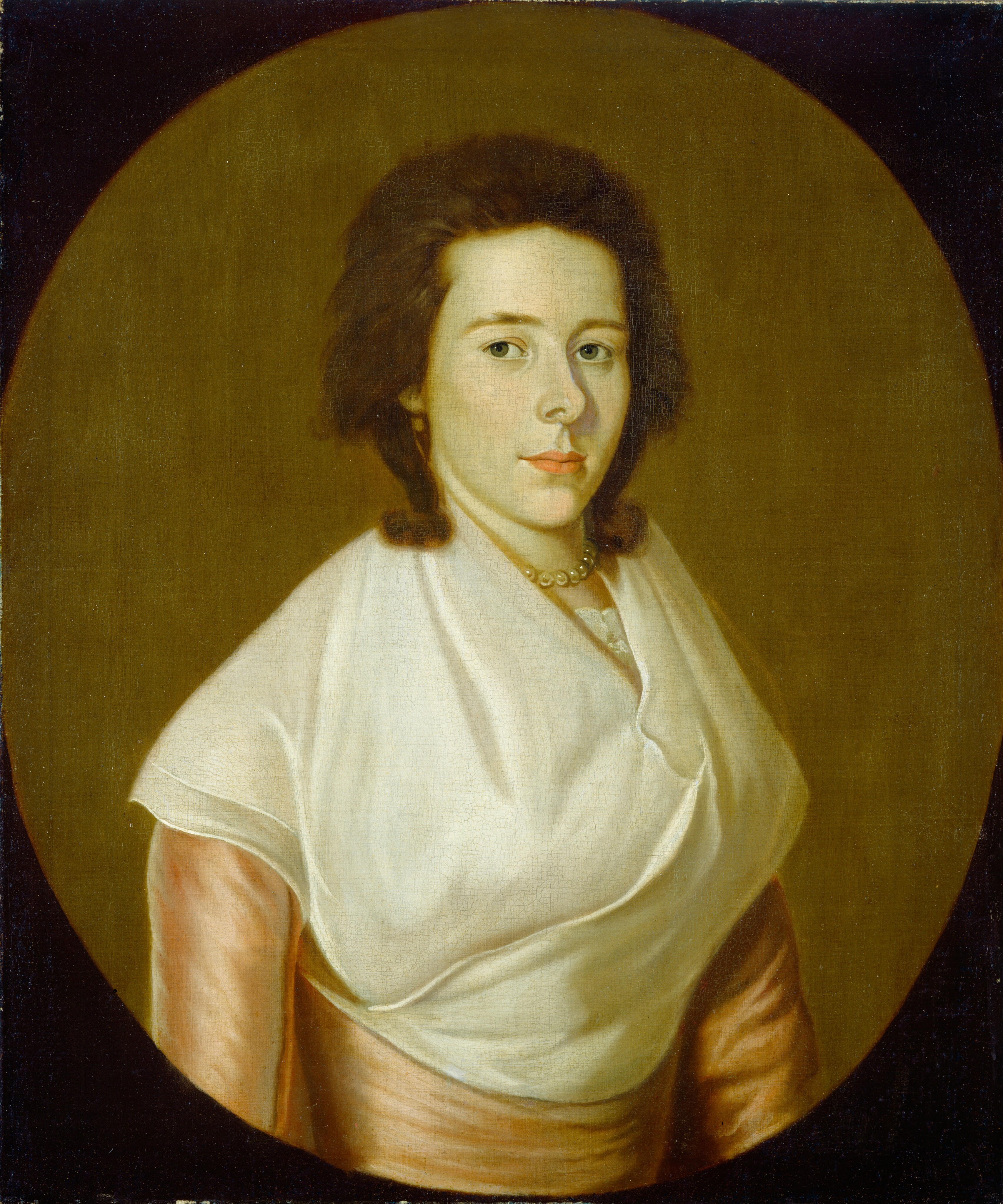 The image shows a woman's upper body. She is looking directly at the viewer with a calm expression. The woman has fair skin, dark brown hair styled away from her face, blue eyes, and delicate features. She is wearing a soft pink dress with a white shawl draped over her shoulders and a pearl necklace. The background is plain and brown.