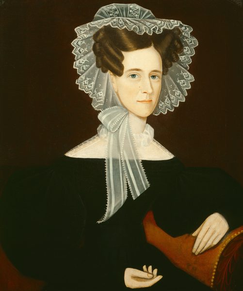The image shows a portrait of a woman. She is seated with her body turned slightly to her left, her right arm resting on a reddish-brown plush armchair. Her face is turned towards the viewer, with calm blue eyes, a faint smile, and clearly defined features. Her hair is styled in elaborate curls framing her face, and she wears a white, lace-edged bonnet with a wide ribbon tied beneath her chin. She is dressed in a dark dress with a high neckline and wide, puffed sleeves. The background is a plain, dark color.